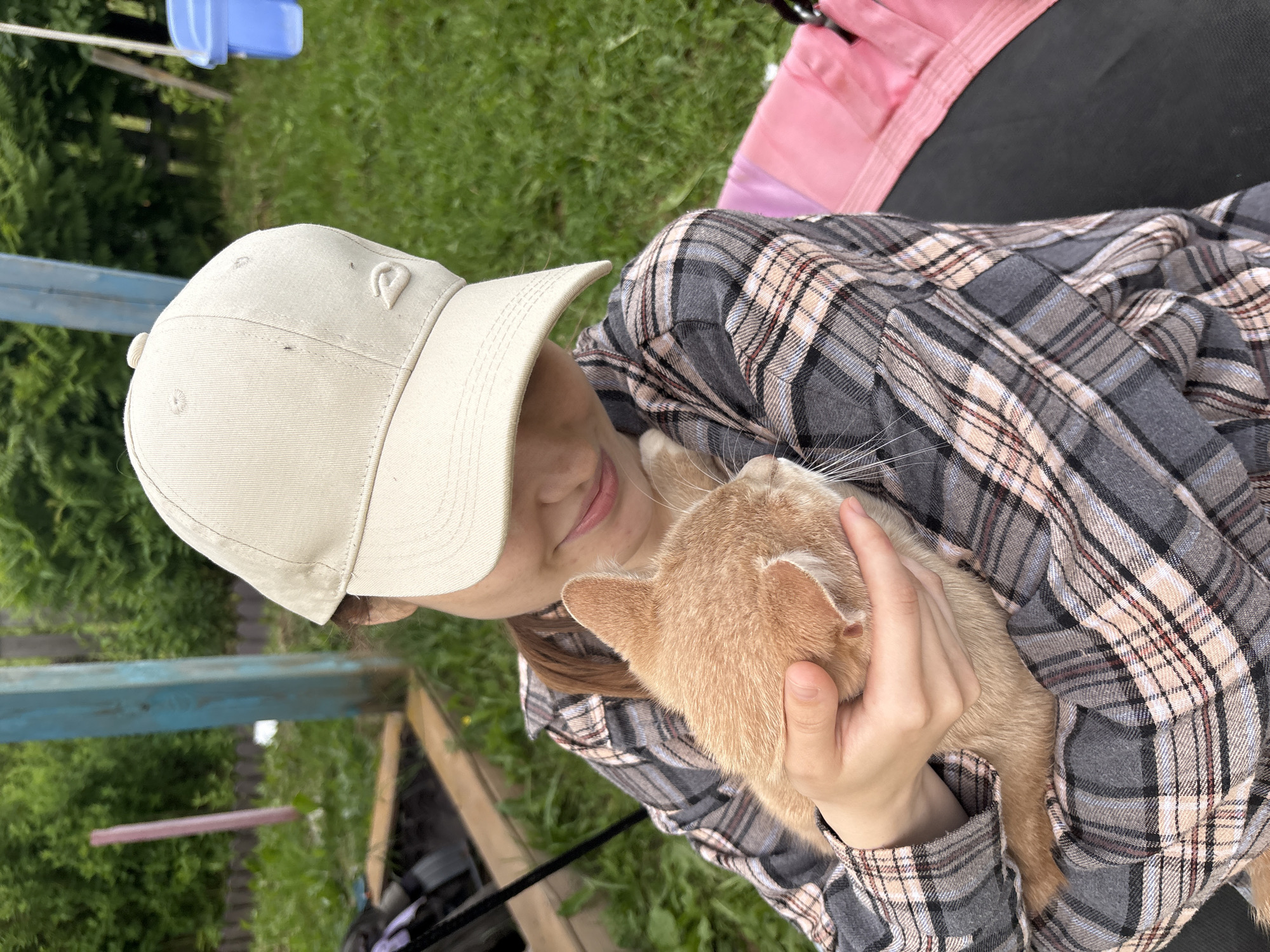 An adult female wearing a beige baseball cap and a grey, pink, and white plaid long-sleeved shirt is holding a ginger cat. The woman is looking down at the cat with a gentle expression. The cat is nestled in her arms, facing away from the camera. The background appears to be an outdoor setting with green grass, bushes, and a wooden fence. There is a blue plastic bin and part of a swing set visible in the background. The image was taken in the city of Kumyon, Russia.