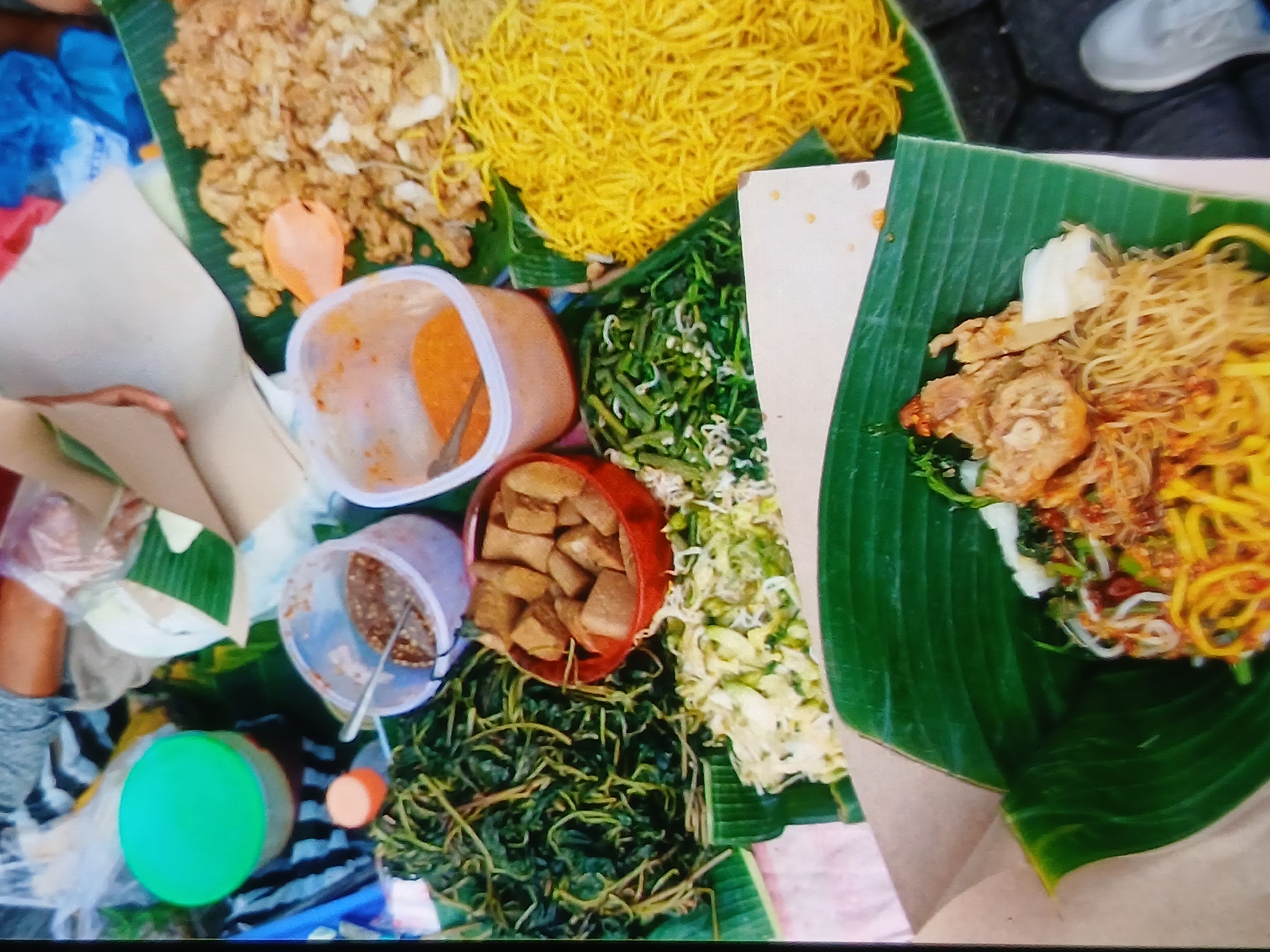 The image depicts a street food stall in Setia Bakti, Indonesia. A variety of Indonesian dishes are displayed on banana leaves. The dishes include fried noodles, stir-fried vegetables, fried tofu, and what appears to be chicken or meat. There are also two containers of sauces or dips. In the background, people's hands and parts of their clothing are visible, suggesting an active vending scene. The setting appears to be outdoors, with a paved ground visible at the bottom right.