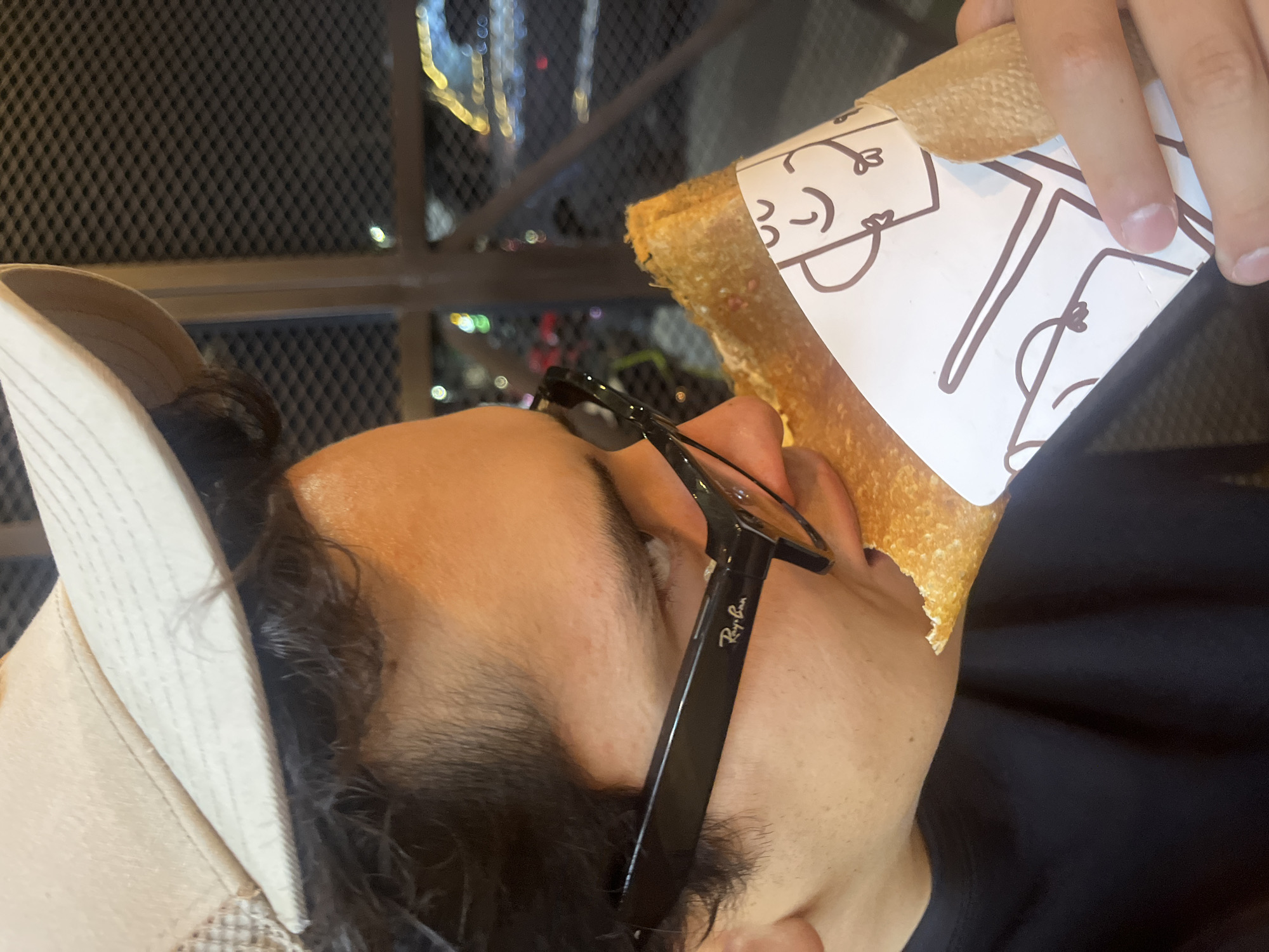 A person is shown in a close-up view, apparently taking a bite out of a crepe. The crepe is wrapped in white paper with drawn faces on it. The individual wears a light-colored cap and black-framed Ray-Ban sunglasses. The background includes an outdoor setting at night, with blurred lights suggesting a city environment. The scene appears to be located in Puebla, Mexico.