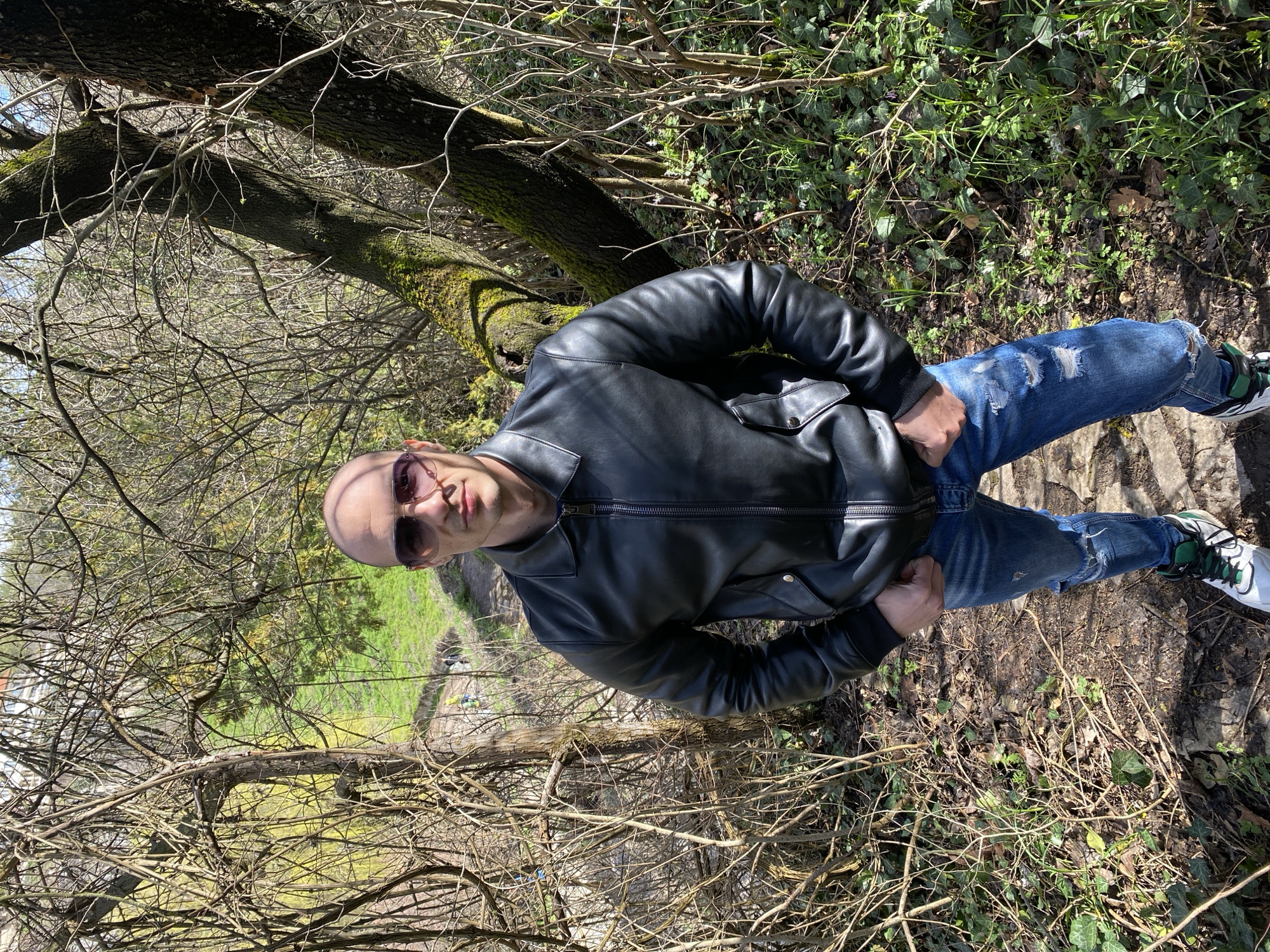A single male subject is depicted standing on a dirt path within a wooded or park-like area in Pleven, Bulgaria. The subject is bald, wearing dark, possibly purple-tinted, aviator-style sunglasses. He is dressed in a black leather bomber jacket with a zippered front, blue distressed denim jeans featuring multiple tears, and white, green, and black athletic shoes. His hands are partially obscured, forming fists at hip level. The immediate environment is characterized by dense, mostly bare deciduous tree branches and shrubs, indicating a late winter or early spring season. A prominent tree trunk covered in green moss is situated to the subject's right. The ground features a mixture of exposed dirt, fallen leaves, and patches of green ground cover, including ivy. In the mid-ground, a downward-sloping grassy area transitions into a darker, possibly excavated section where faint human figures and a yellow element are discernible. Residential structures are visible on the distant horizon above the treeline. The scene is illuminated by strong, direct sunlight, creating distinct shadows.