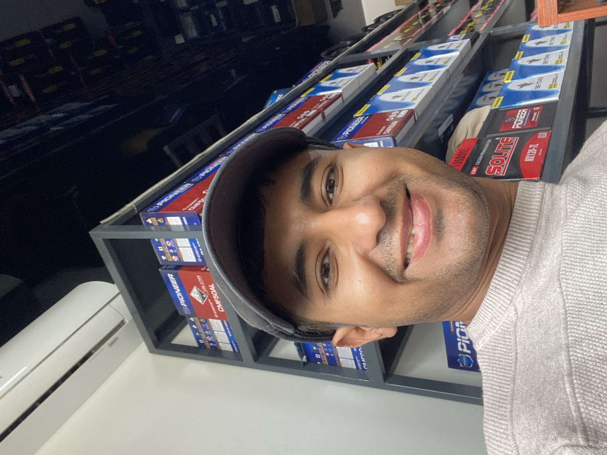 The image shows a man in a gray hat and sweater standing in what appears to be a retail store. Behind him is a shelving unit filled with car batteries and related products from brands like Pioneer, Solite, and Beyonx. The labels on the products show information such as "CMF50AL", "CMF CALCIUM PREMIUM BATTERY", and "NX120-7L". An air conditioning unit is visible on the left side of the frame. The specific location is not discernible from the image.