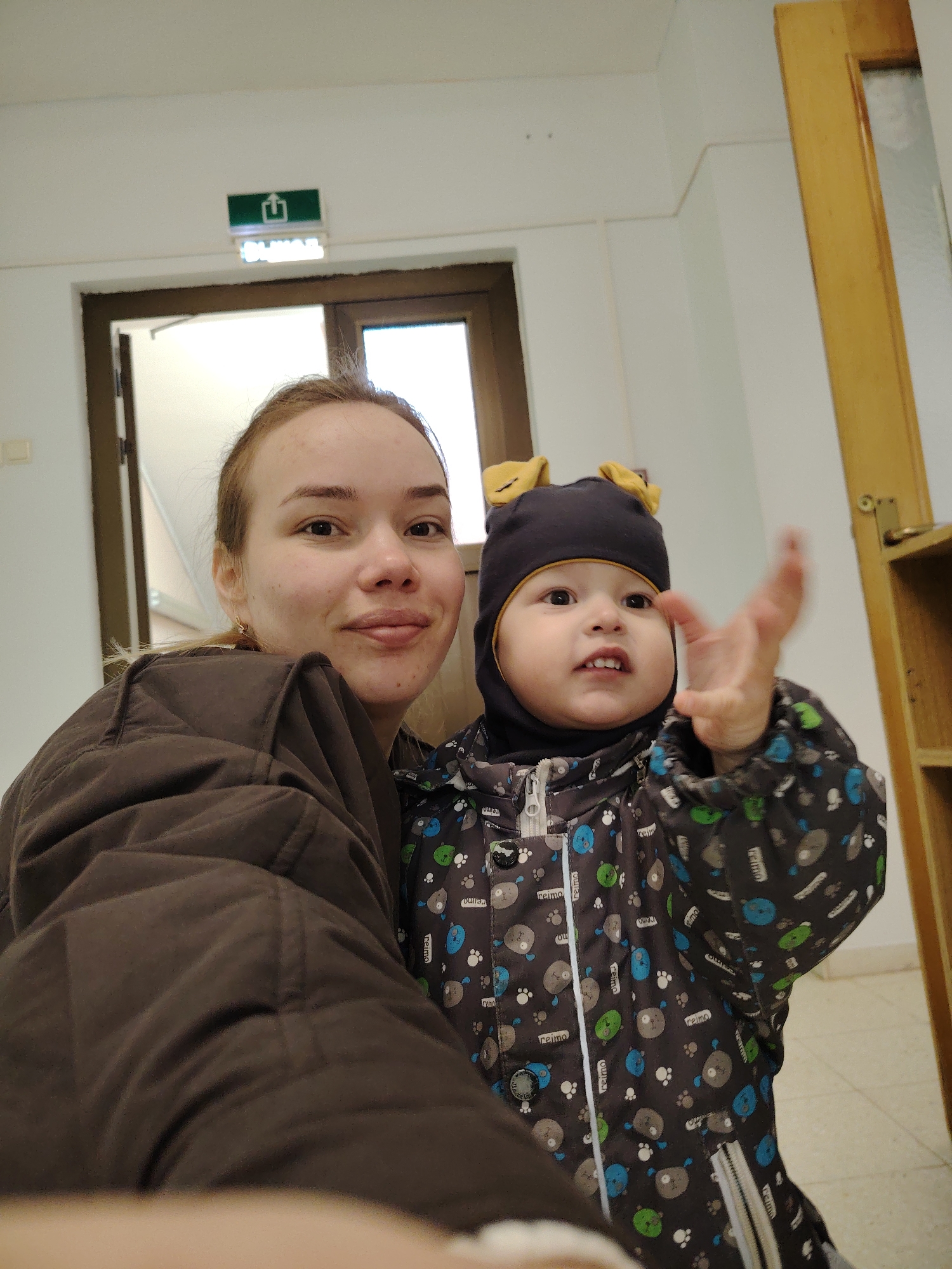 The image is a selfie taken indoors. A young woman with light brown hair and fair skin is visible, wearing a brown jacket. Next to her is a young child wearing a dark-colored winter suit with a pattern of cartoon bear faces. The child also wears a matching hat with small yellow ears. The child is holding their right hand up, fingers spread.
In the background, a doorframe with a green exit sign above is visible, suggesting a public building or apartment complex. A light-colored wall and a wooden structure are also present.
Based on the "Reimo" brand on the child's suit and the exit sign in Cyrillic, the location is most likely in Russia. The suit brand appears to be available in Markovskii, Russia, thus the photo may have been taken there.