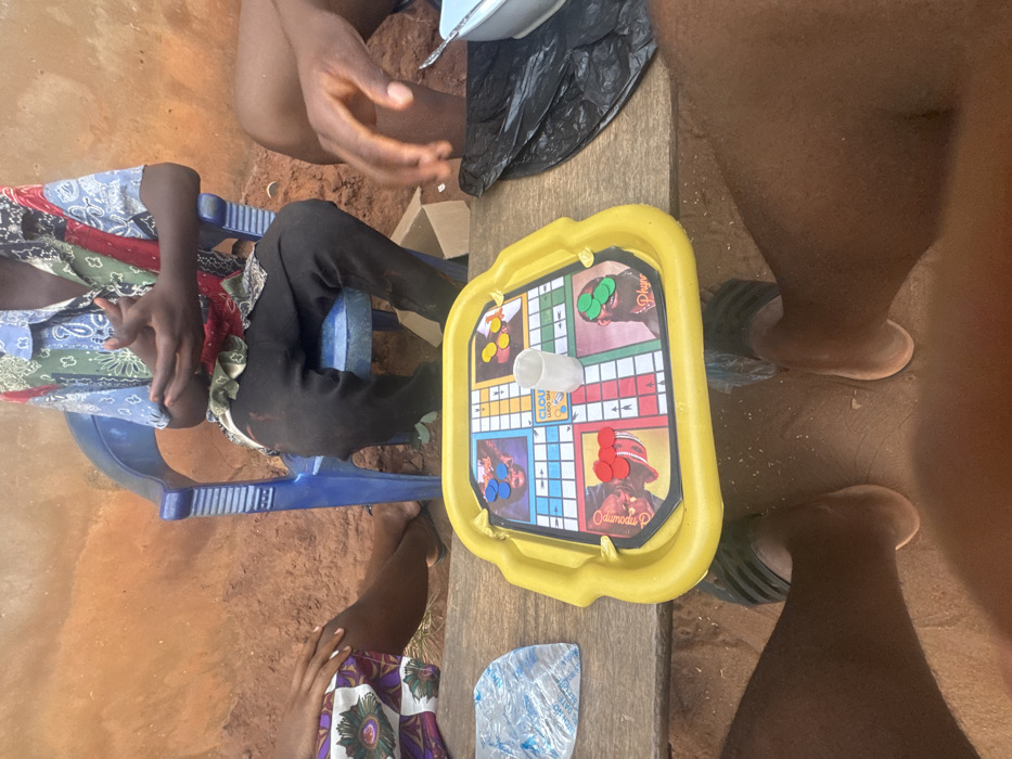 Four people gather around a yellow board game, likely Ludo, placed on a wooden surface. The game features faces of what appears to be musicians. A person wearing a colorful shirt sits on a blue plastic chair, while another wears a patterned skirt. A white dice cup sits in the center of the board, surrounded by game pieces. The scene is shot at an awkward angle, possibly from above, and includes partially visible legs and feet of the individuals playing.