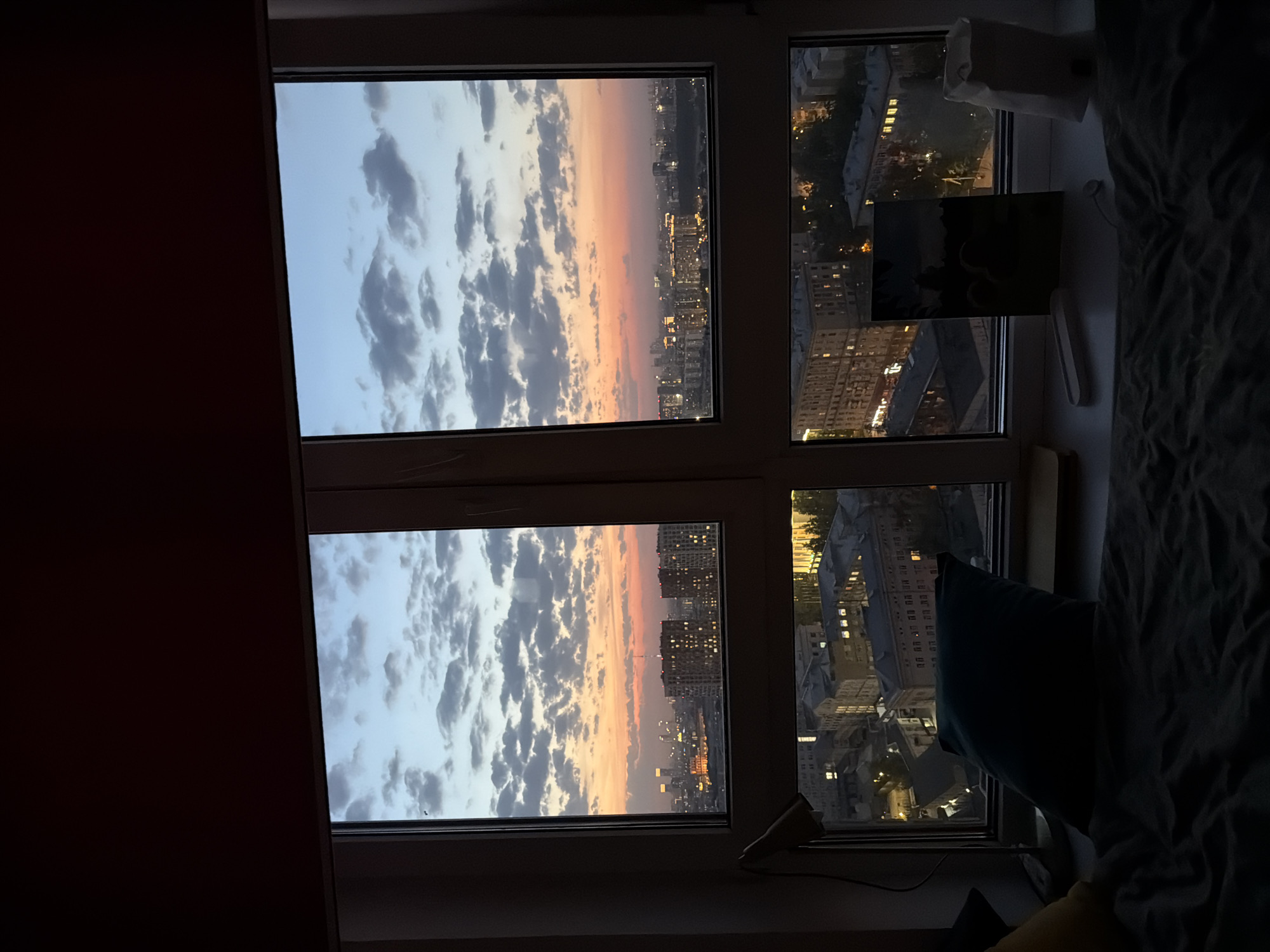 The photograph shows a view through a multi-pane window at dusk or dawn. The sky is visible through the two panes on the left, displaying a range of colors from blue to orange, with textured clouds. A cityscape is seen through the window panes on the right, with lit buildings indicating it is either early morning or late evening. The window is situated in a room, with part of the room visible on the right side, including bedding, pillows, and objects on the windowsill. The scene appears to be in a residential building in an urban area.