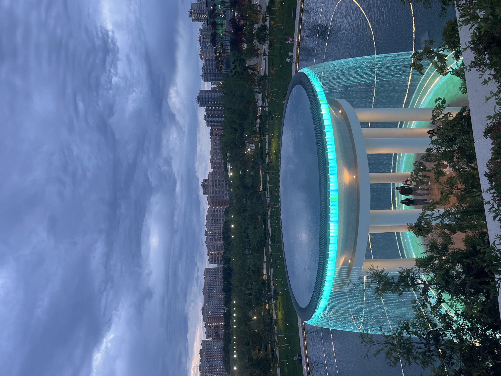 This image captures an evening scene in a modern urban park located in Krasnodar, Russia. The central subject is a large, circular fountain complex. Its upper basin features a rim illuminated with a bright turquoise-cyan light, reflecting softly on the water's surface. Beneath this, a continuous sheet of water cascades downwards, also glowing with the same vibrant turquoise light, supported by several prominent white architectural pillars. Further down, golden-yellow illuminated water jets arc gracefully over a lower water level, creating additional visual interest.
Several people are visible enjoying the fountain area, appearing as silhouettes against the illuminated structures, some standing near the pillars, others observing from nearby paths. Beyond the fountain, a well-maintained park extends into the distance, characterized by green lawns, numerous trees, and subtle pathway lighting. A long line of multi-story residential buildings forms the distant backdrop, their windows showing scattered lights, suggesting urban occupancy.
The time of day is clearly evening or late dusk, as indicated by the prominent artificial lighting and the darkening, overcast sky with shades of blue and grey clouds. The overall atmosphere is calm and inviting, showcasing a beautifully designed public space. No discernible text is visible within the image.