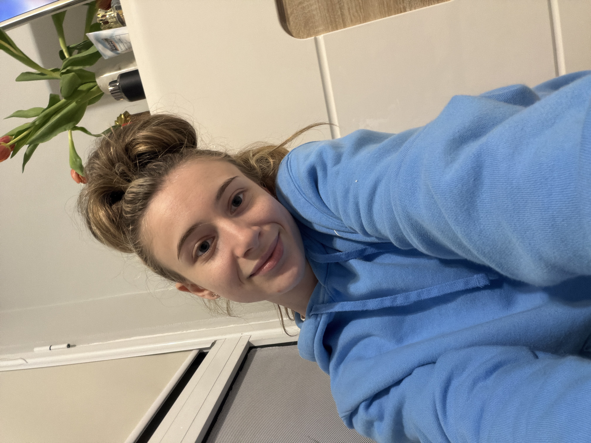 A young woman with blonde hair in a messy bun is pictured indoors, wearing a light blue hooded sweatshirt. She is smiling and looking directly at the camera, which is positioned at a low angle. To her left, there are green tulips in a container, and a white wall is behind them. Below her, there's a window with a screen and a white frame. The location within the city of Plovdiv, Bulgaria cannot be determined from the image.