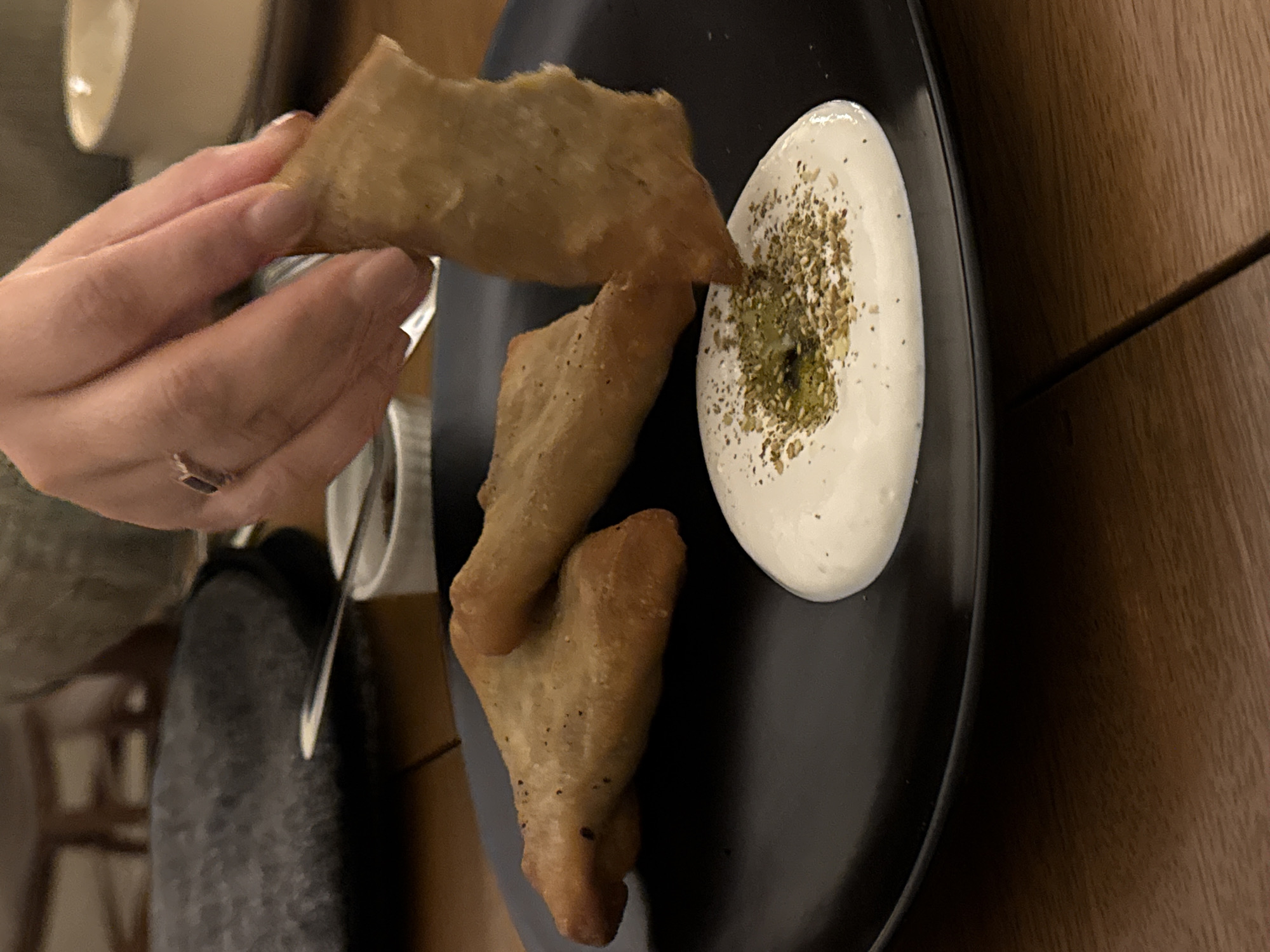 A hand holds a fried triangular pastry over a black plate. The plate contains two more pastries of the same type and a dollop of white sauce sprinkled with green and brown spices. The setting appears to be a restaurant on a wood-grain table. The location is specified as Unknown, Unknown.