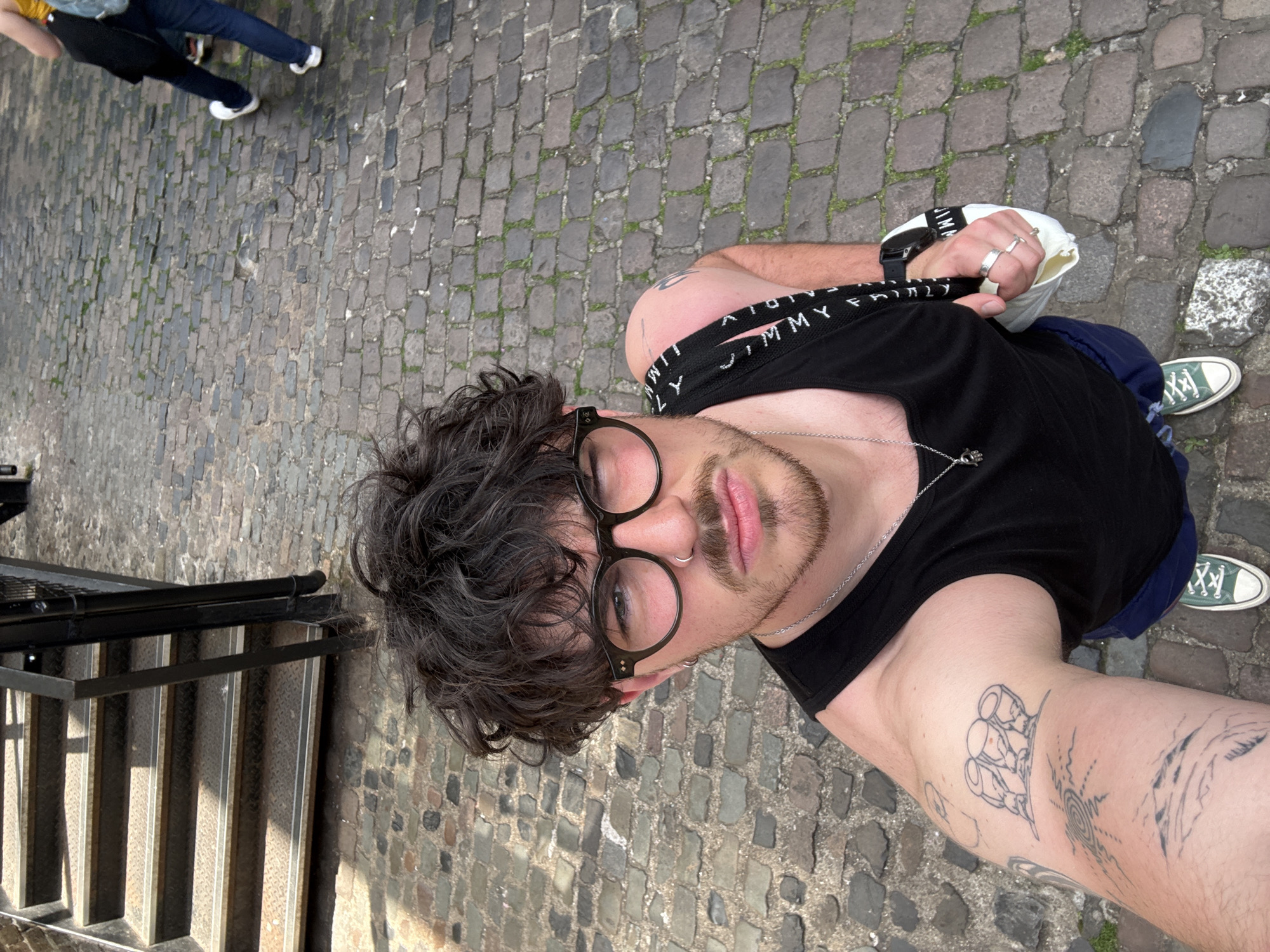 A person is taking a selfie in the Unknown, Unknown, on a street with cobblestone pavement. The person is wearing a black tank top, dark-rimmed glasses, and has curly dark hair. Visible tattoos are on the person's arms. The person is holding a white bag in one hand. To the left of the person are metal stairs. In the background, other people are walking.