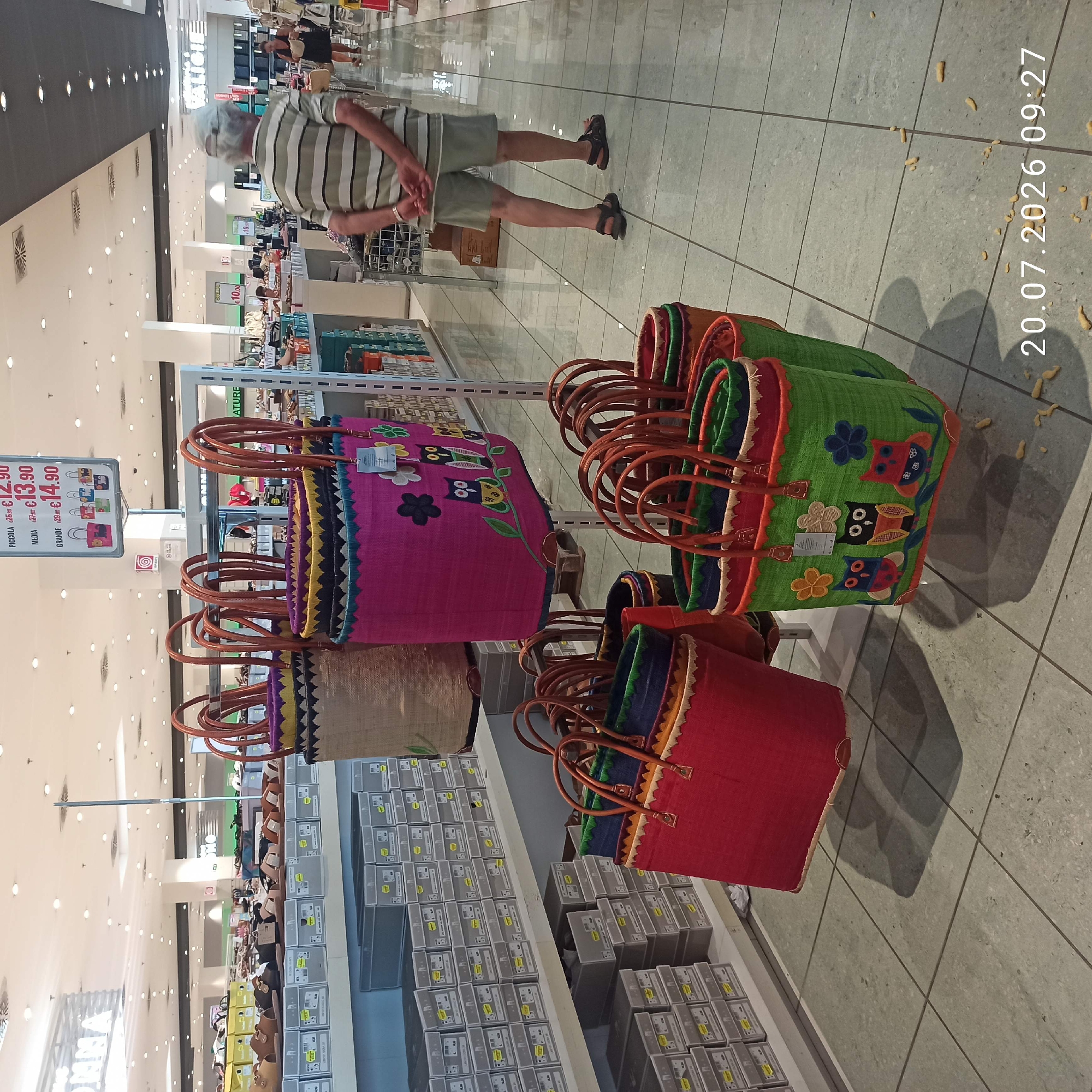 In a retail store in Bari, Italy, the image focuses on a display of woven tote bags. The bags are colorful and feature embroidered designs, including owls and flowers. They are arranged on a multi-tiered stand, with some bags hanging and others sitting on shelves. In the background, rows of shoe boxes are visible on shelves, suggesting the store sells footwear. A man wearing a striped shirt and shorts, with his back to the camera, is standing in the aisle, looking towards the merchandise. Other shoppers and store displays are partially visible in the distance, indicating a busy retail environment. The flooring is tiled, and the ceiling features fluorescent lighting. The timestamp "20.07.2026 09:27" is overlaid on the bottom right corner of the image.