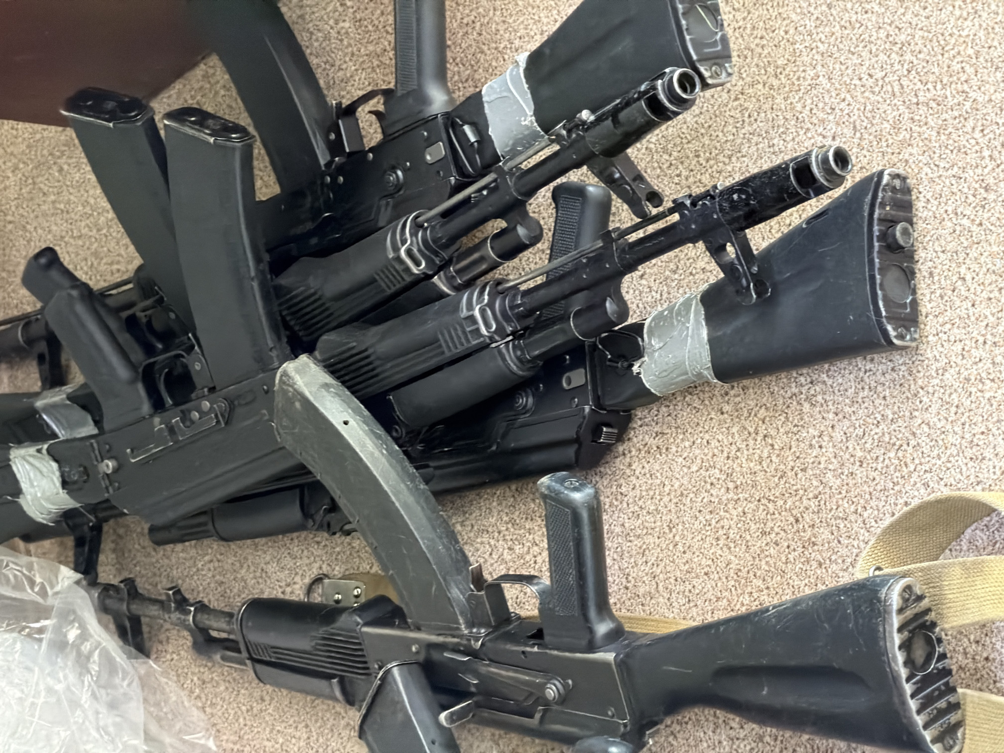 The image shows a collection of black AK-type rifles laid on a light brown carpet or rug. The rifles are positioned at various angles, overlapping each other. The magazines are inserted into the rifles. Some of the rifles have tape around the stocks. A tan-colored strap is partially visible in the lower right corner. There is no indication of people or location, including the city of Забегалово, Russia.