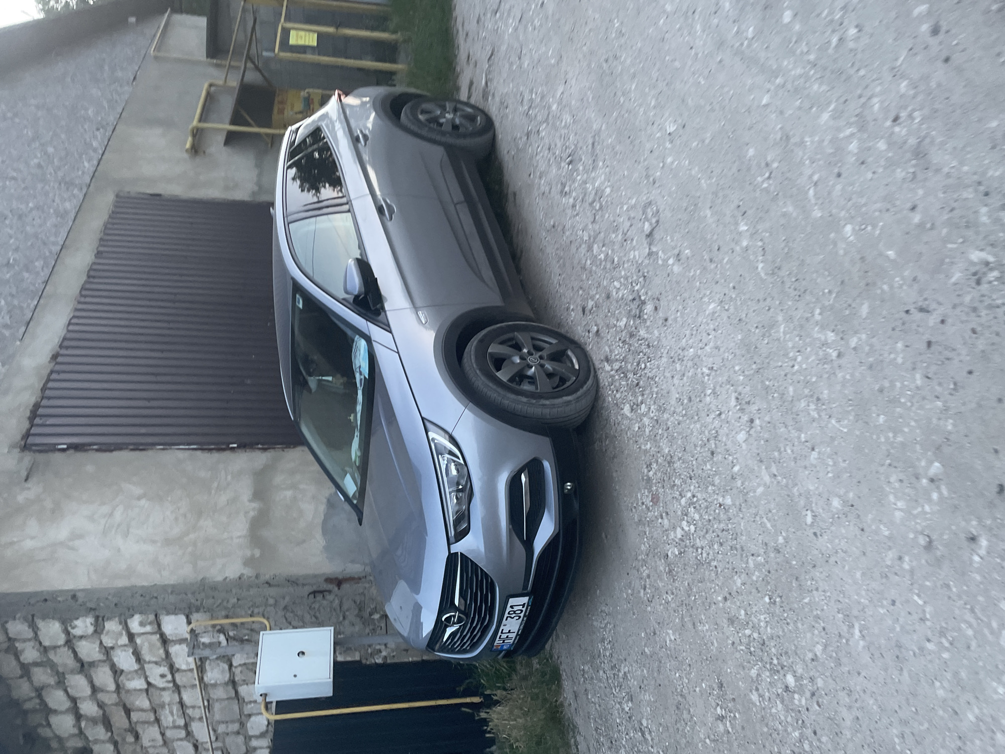 This media file captures a gray Opel Astra parked on a gravel surface. The car is angled, with its front facing towards the right of the frame. The vehicle appears to be in a somewhat utilitarian setting, possibly a rural or industrial area, with a building constructed of brick and concrete visible in the background. A brown corrugated metal door or panel is present on the building's exterior, and a white electrical box with yellow conduits is mounted on the wall.
The lighting suggests it is daytime, though the overall atmosphere is somewhat muted, implying it could be overcast or late afternoon. The car itself is the primary subject, with details like its license plate clearly visible. The license plate displays the text "HFF 381". The scene is static, with no people or apparent activity taking place. The car's wheels are dark gray alloy rims. The backdrop includes elements of urban or semi-urban infrastructure, including scaffolding-like structures and what appear to be utility lines. The ground is primarily composed of gravel and dirt.
