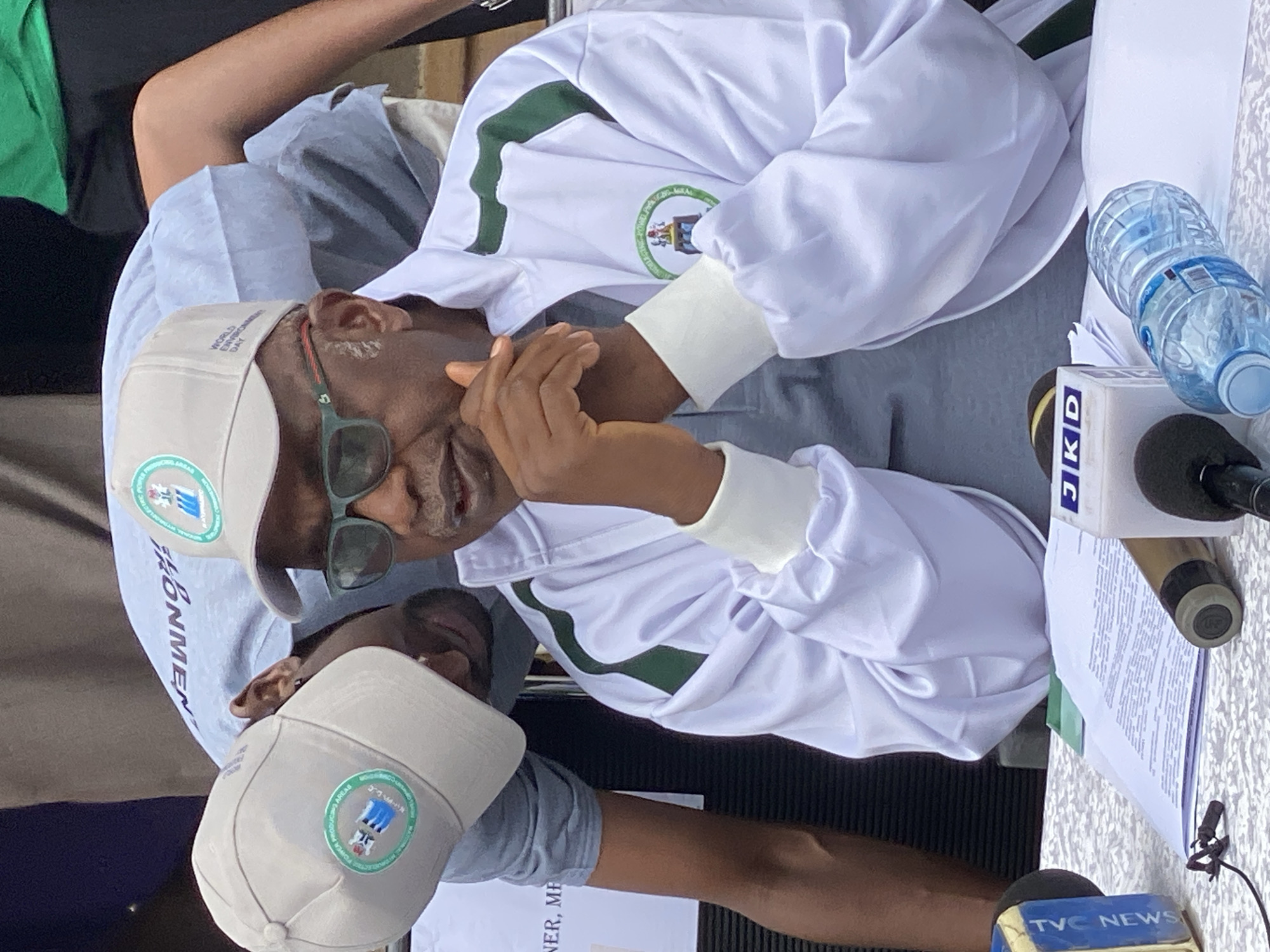 The image, taken in Shaba, Nigeria, captures what appears to be a press conference or official event. The central figure is an older man, seen from the chest up, wearing a white jacket with green stripes and a light-colored cap. He also sports dark sunglasses and has his hands near his face, suggesting he is speaking or listening intently. His cap bears the text "WORLD ENVIRONMENT DAY" and a circular logo that reads "NATIONAL ELECTRIC POWER PRODUCING AREAS." The white jacket also features a similar circular logo.
Partially visible below him is another individual wearing a similar light-colored cap, which also displays the "NATIONAL ELECTRIC POWER PRODUCING AREAS" logo.
The scene includes a table with papers, one of which shows visible text like "HONMEN" and "NER, ME." In the foreground, microphones from news organizations "JKD" and "TVC NEWS" are present, confirming the media context of the event. A clear plastic water bottle is also on the table. The lighting suggests it is daytime, and the setting appears to be an outdoor or semi-enclosed public space.