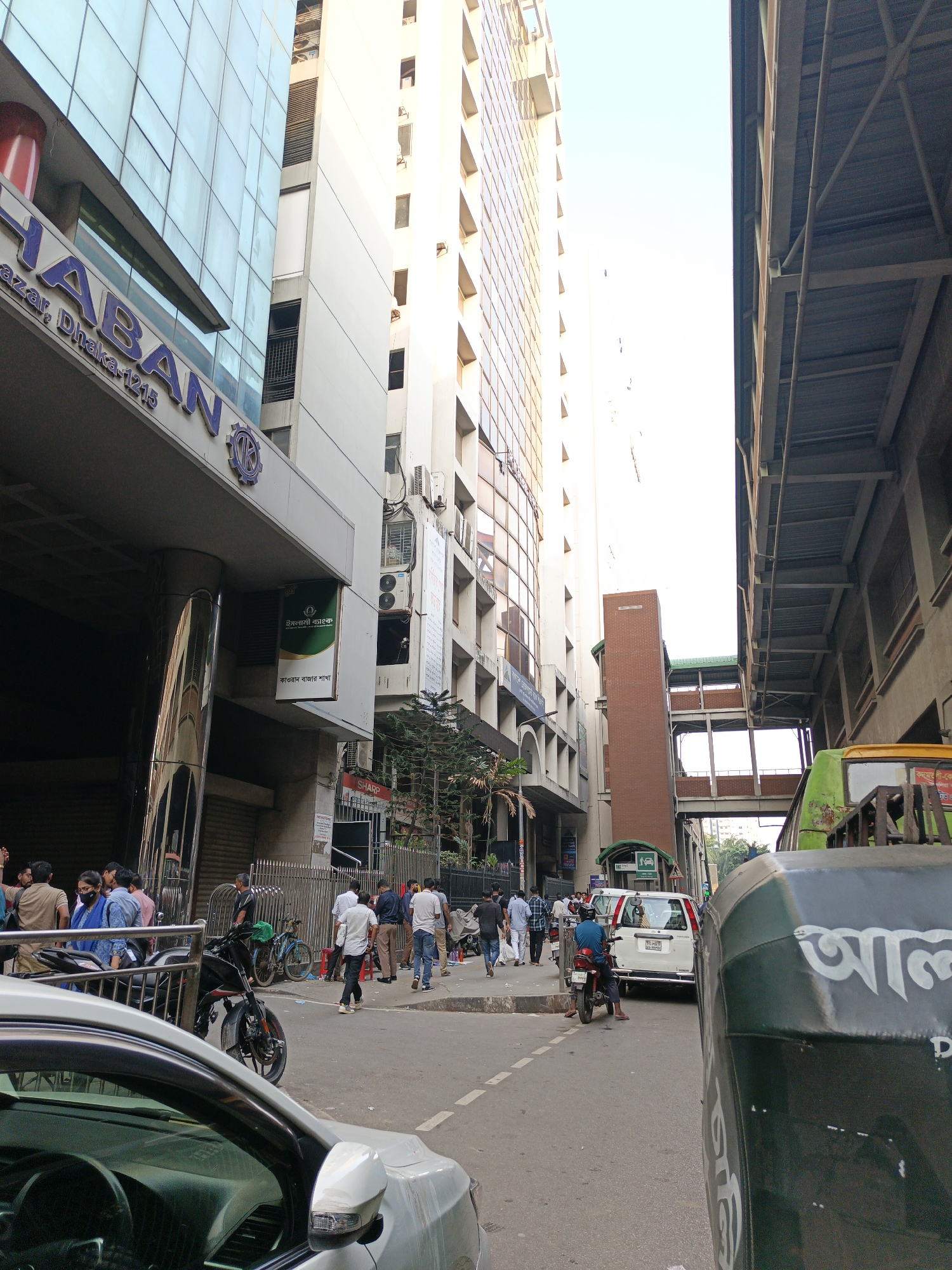 The image captures a street scene in Dhaka, Bangladesh. On the left is a tall building with glass and concrete facade, marked with signage for "HABAN" and "Islami Bank". People are gathered on the sidewalk near the building, some standing and talking, others walking. Bicycles and motorcycles are parked along the curb, and parts of a white car are visible in the foreground.
The street is lined with vehicles, including a white van and a green bus. In the background, a pedestrian overpass can be seen, constructed with metal beams and concrete supports. The sky is overcast, casting a soft light on the scene. The overall impression is of a busy commercial area in Dhaka.