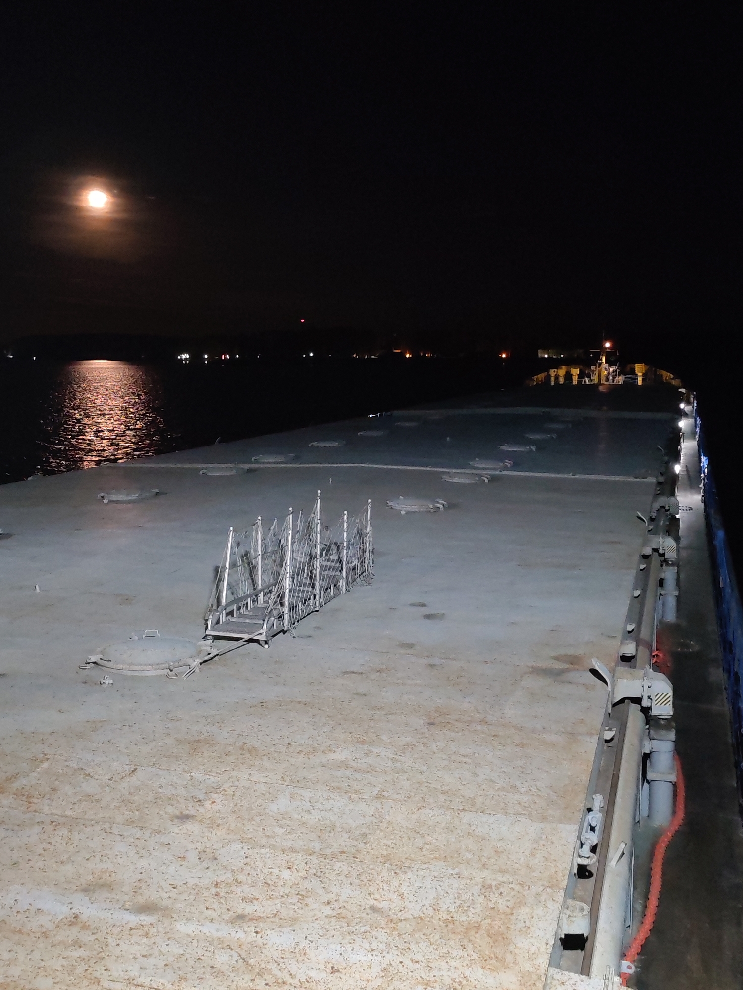 The image depicts the deck of a ship at night. The deck is flat and gray, with a raised metal structure in the center of the frame. A set of stairs with metal railings is present on the deck. The deck is lit by lights on the side of the ship and the faint light of a full moon in the sky. In the distance, there are lights on the shoreline. The water is dark and reflects the moonlight.