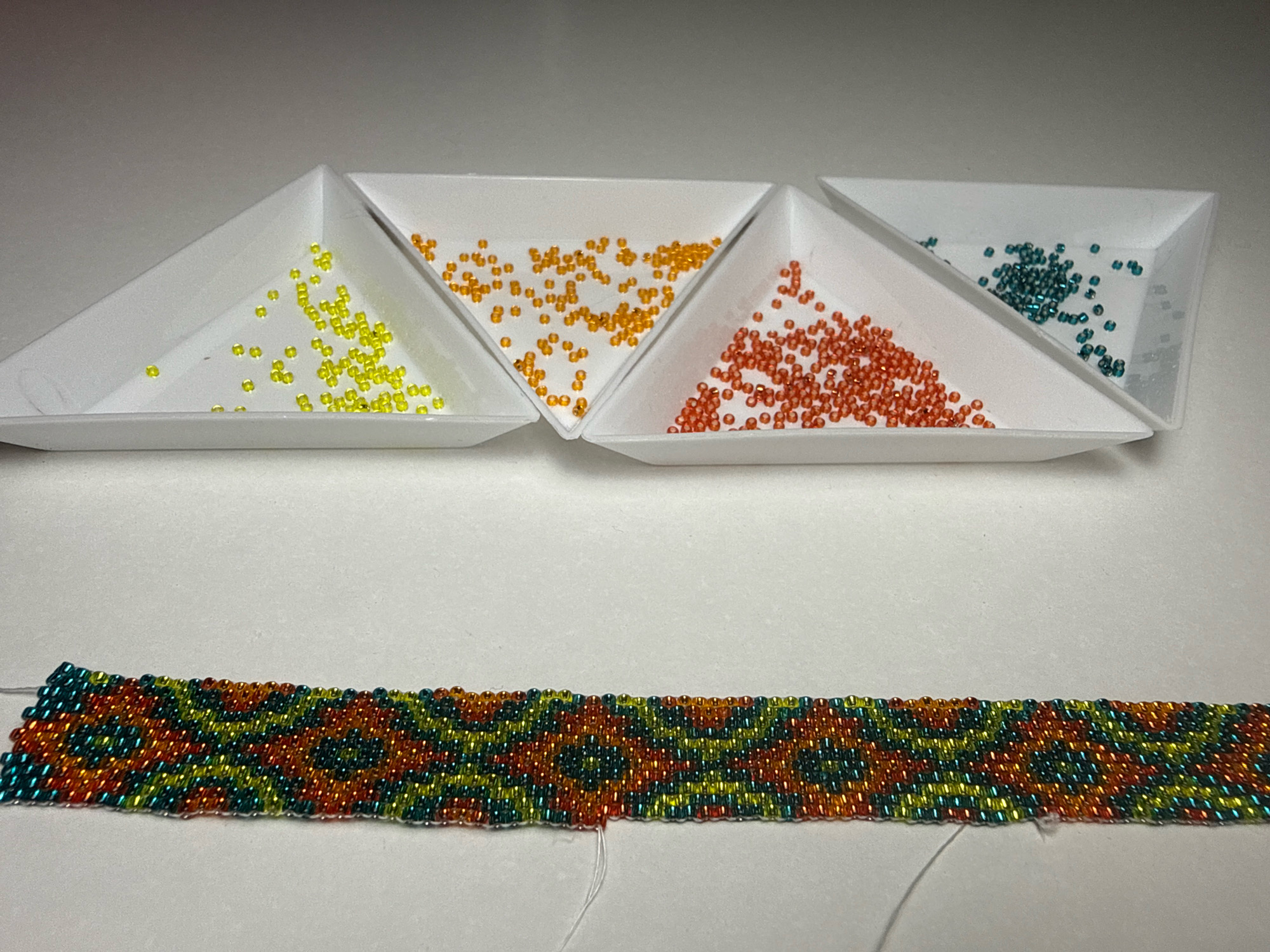 This image shows a close-up, overhead view of four triangular white trays filled with colorful seed beads, arranged diagonally from left to right. The trays contain yellow, orange, reddish-orange, and teal beads respectively. In front of the trays, a beaded bracelet is laid out horizontally. The bracelet exhibits a geometric pattern in teal, yellow, orange, and reddish-orange beads. A white thread is visible extending from the left end of the bracelet. The background is a plain, off-white surface. There are no elements in the image that indicate a location in Kazan, Russia.