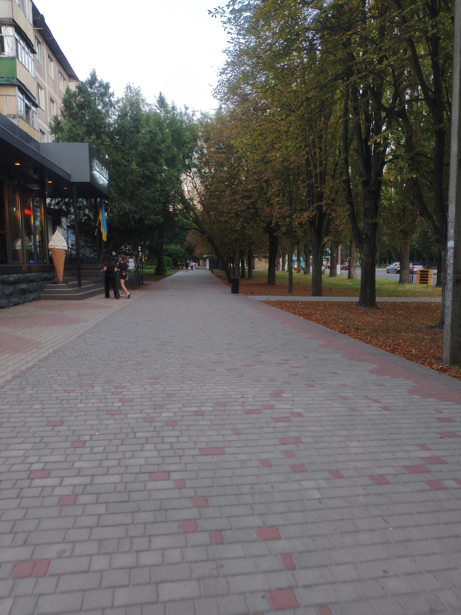 The image shows a paved walkway in Kremenchuk, Ukraine, adjacent to a building and a street lined with trees. On the left, a building with balconies and windows is partially visible. In front of the building, a large decorative ice cream cone stands near the entrance. Two individuals are walking on the walkway. The walkway itself is paved with grey and red rectangular bricks arranged in a pattern. On the right side of the walkway, trees with autumn foliage line the path, and beyond them, a street with passing cars is visible. The sky is overcast.