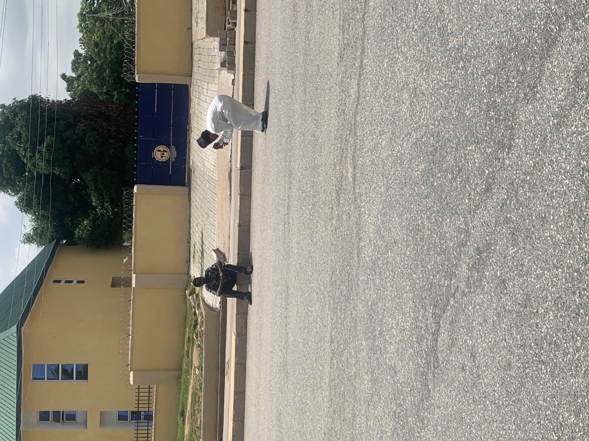 In the city of Jalingo, Nigeria, on a paved road in front of a yellow building with a green roof, two men are visible. The building has several windows and is enclosed by a yellow wall topped with barbed wire. A large blue gate with a circular emblem is centered in the wall.
To the left, a man sits on the curb, facing away from the camera. He is wearing a dark, patterned shirt and dark pants.
To the right, a man in white attire, including a cap, is bending down and appears to be taking a photograph with a smartphone. The area around the building suggests a residential or administrative compound. The sky is overcast, indicating a daytime scene.