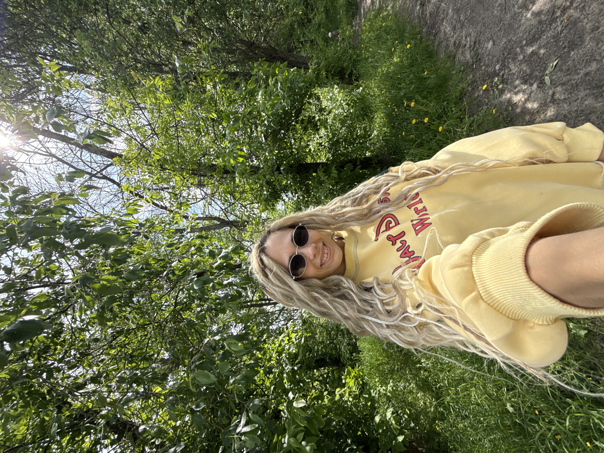 The image is a selfie of a woman with long blonde hair and sunglasses, wearing a yellow sweatshirt with red text. She is smiling at the camera. The background consists of lush green trees and grass, suggesting a park or wooded area. A dirt path is visible in the upper right corner. The lighting suggests a sunny day. No specific landmarks or signage are visible that could verify the location as Rusanovo, Russia.