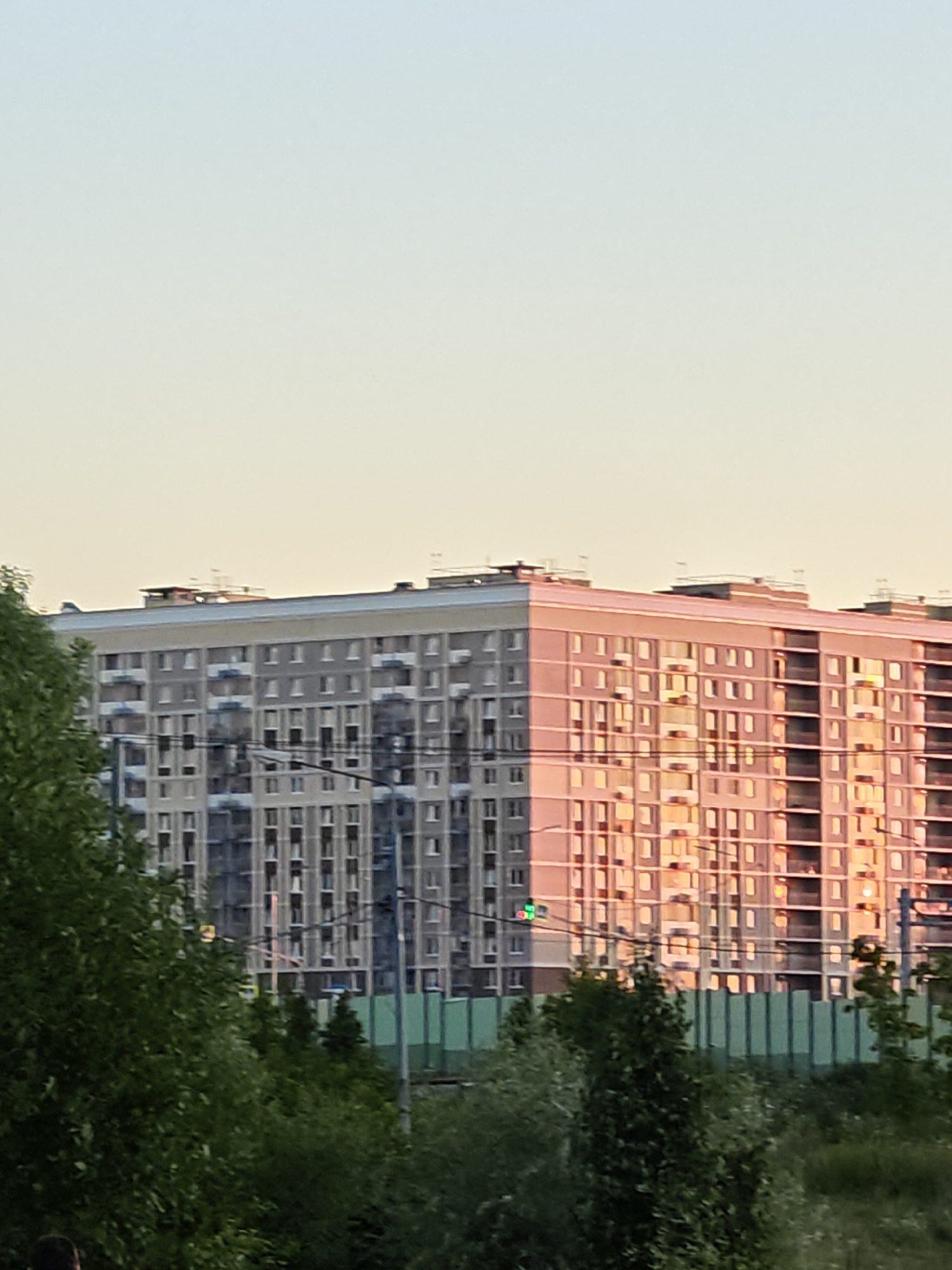 The image captures a large, modern apartment building on a clear day. The building features a repeating pattern of windows and balconies, with one side of the building bathed in the warm glow of the setting sun, creating a striking contrast with the cooler-toned side. In the foreground, lush green trees partially obscure the view of the lower floors. A traffic light with a green signal is visible near the building. In the distance, a green fence runs horizontally across the frame. The sky above is a pale gradient of yellow and blue, characteristic of dusk. The overall scene suggests an urban residential environment in Нефтемаш Снт, Russia.