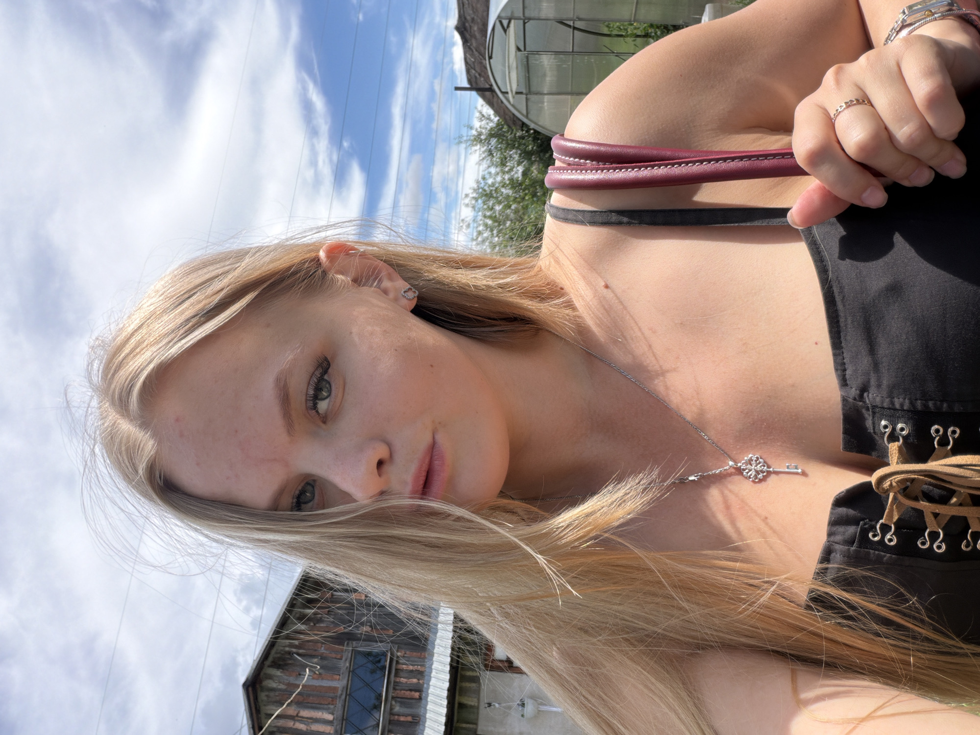 A young woman with blonde hair stands outdoors, holding a purse. She is wearing a black top with laces and a key-shaped pendant necklace. In the background, a wooden building and a greenhouse are visible under a cloudy sky. The location is the city of Chaikovsky, Russia.