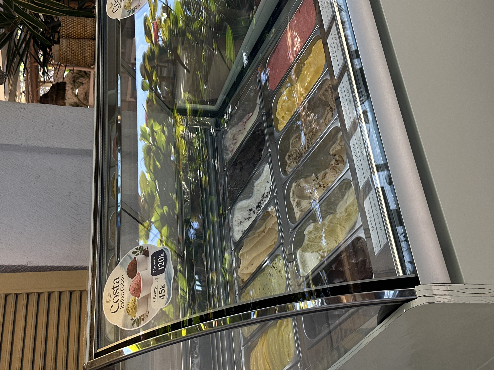An outdoor gelato display case is shown. The display case is filled with various flavors of gelato in metal pans. A sign for "Costa Italian Gelato" is visible on the front of the display case, advertising prices for one and three scoops. The display case is in front of a building with a slatted shutter and a textured wall. Green foliage is visible behind the display case, suggesting an outdoor or semi-outdoor setting. The overall impression is that of an ice cream shop in a tropical or warm climate.