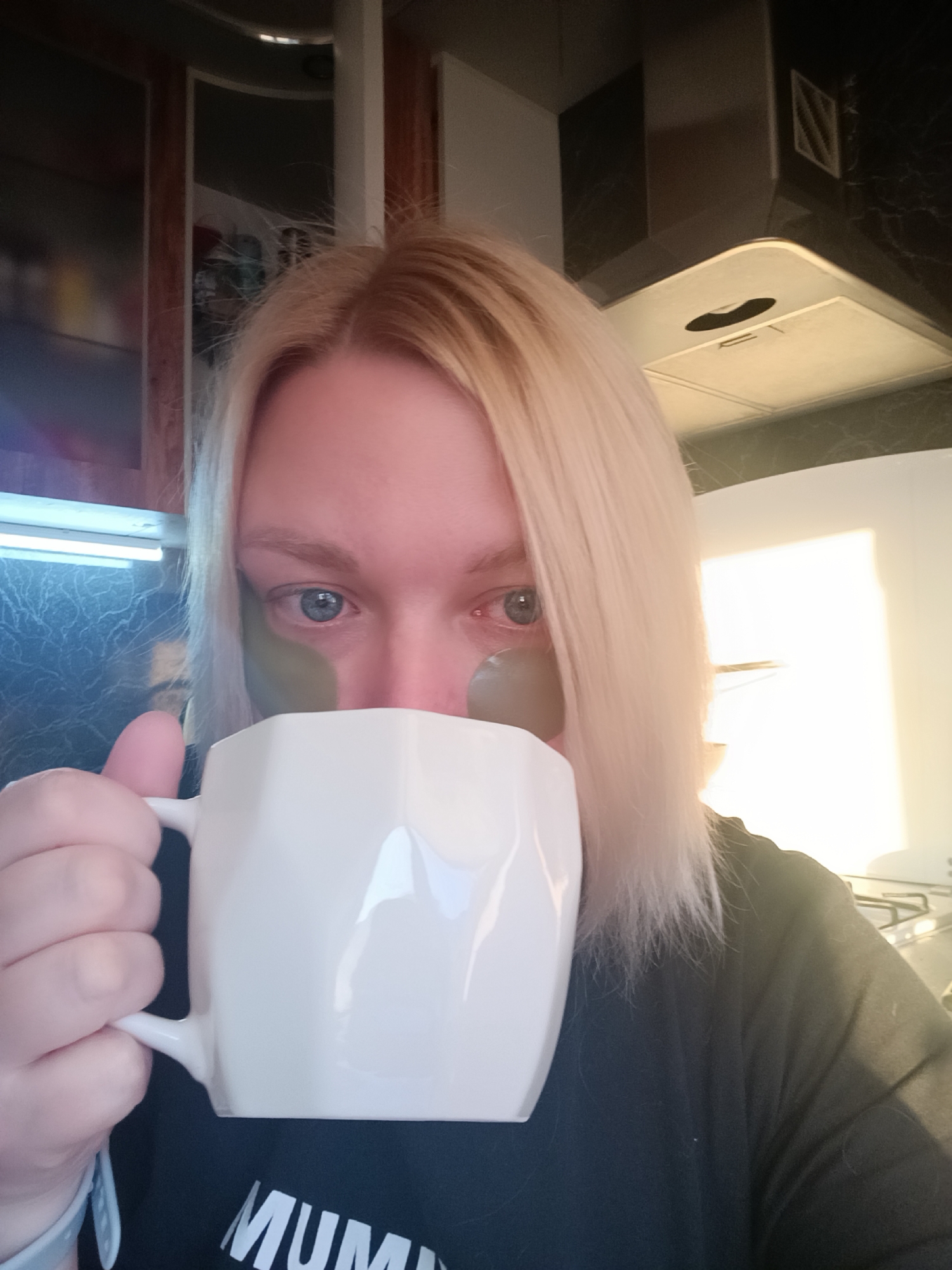 A woman with blonde hair is holding a white mug, partially obscuring her face. She has green eye patches under her eyes. The mug is angular in design. She is wearing a black t-shirt. The background shows kitchen cabinets, a range hood, and a light fixture. It is unclear if this image was taken in the city of Chaikovsky, Russia.