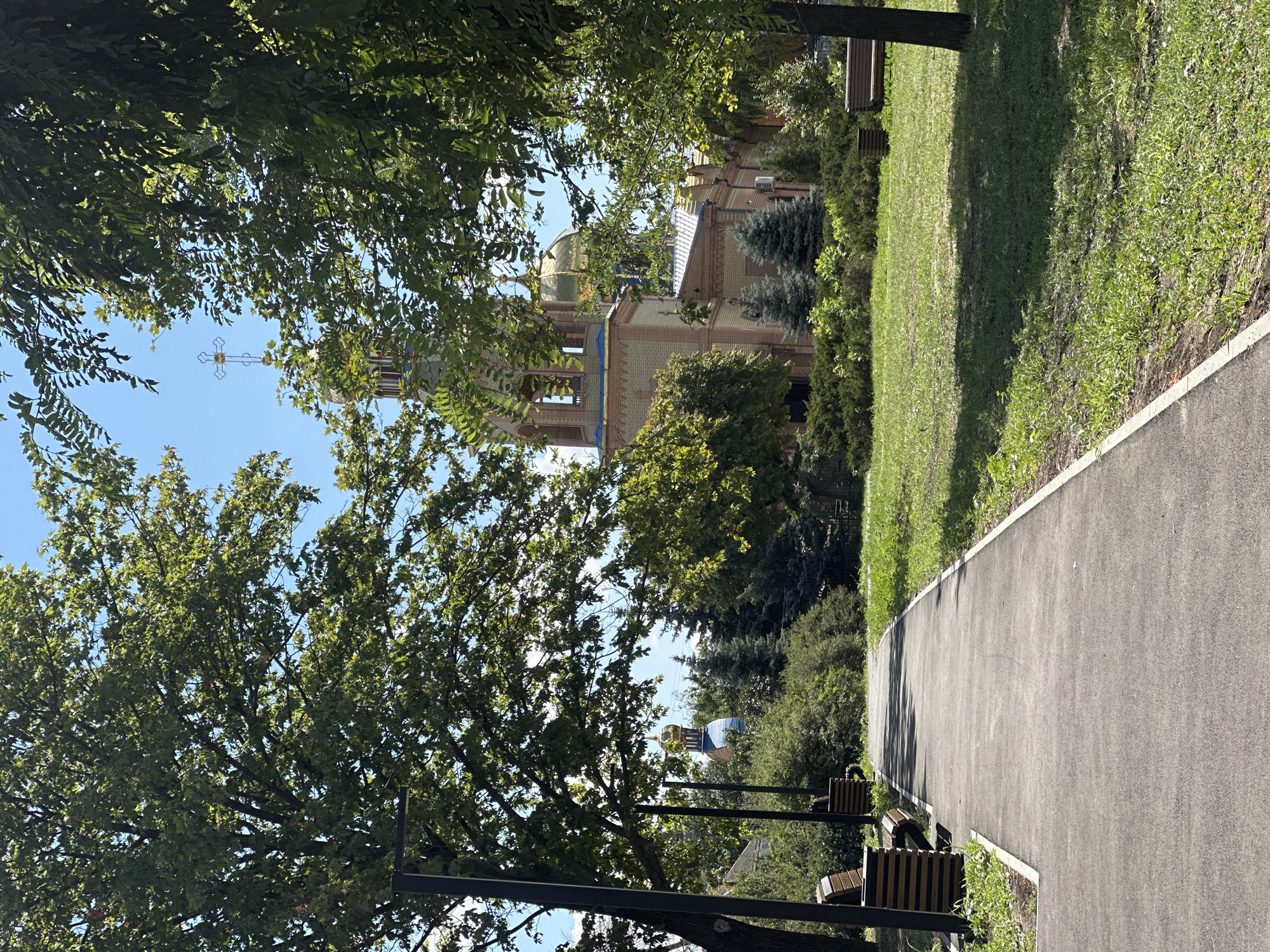 The image captures a park pathway leading towards a church in Zhovte, Ukraine. The pathway is paved with asphalt and bordered by a strip of grass and concrete. On the left side of the pathway, there are two benches. The park features lush green grass and trees, with sunlight filtering through the leaves, creating dappled shadows on the ground.
In the background, a church with a prominent golden dome and a bell tower is visible. The church's architecture suggests it is an Orthodox Christian church, characterized by its religious iconography and design elements common in Eastern Europe. The church is surrounded by trees and greenery, with another building with a blue dome visible in the distance to the left. The sky is clear and blue, indicating a sunny day. The overall scene is peaceful and serene, typical of a public park.