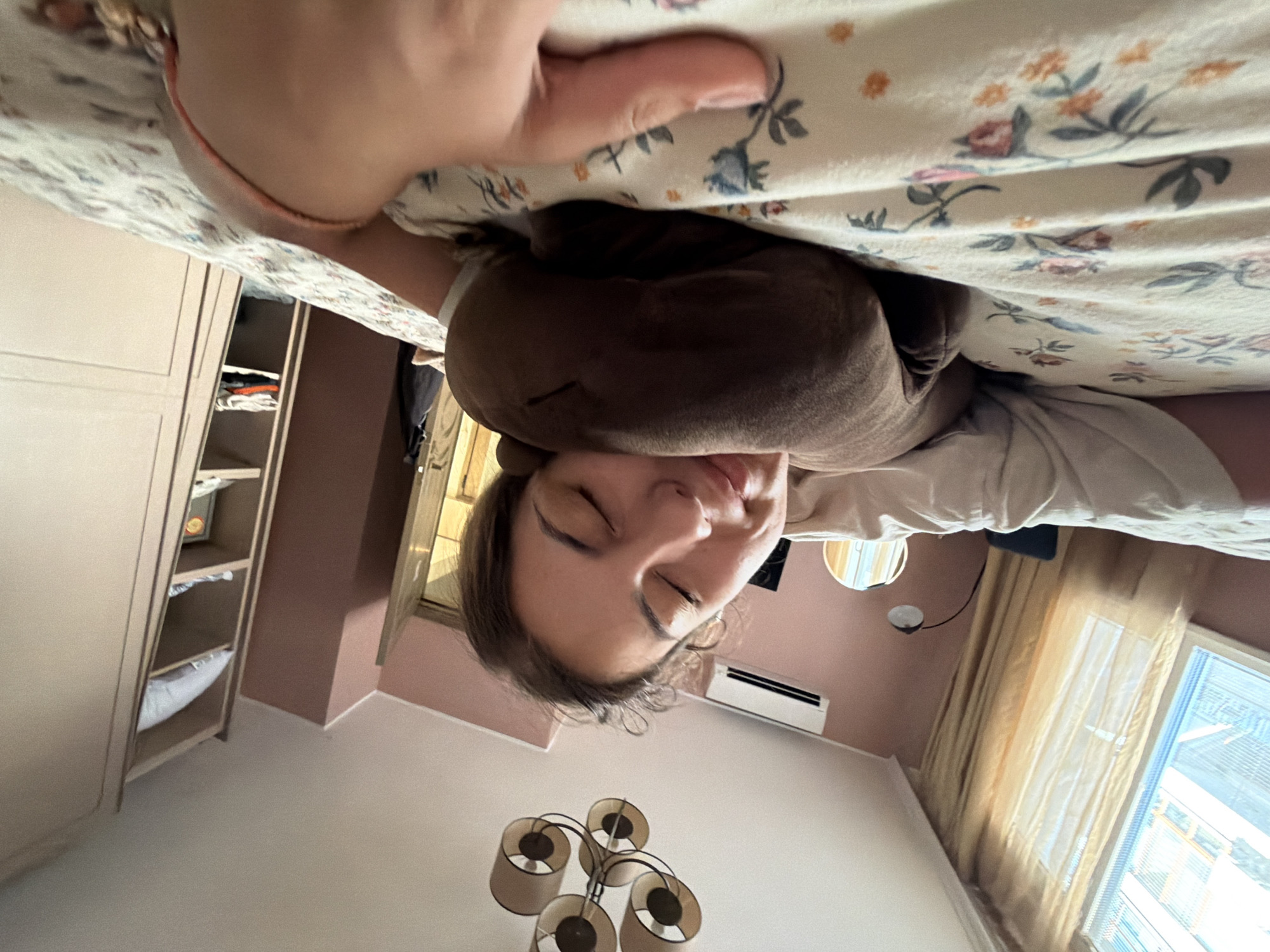 A young woman is sleeping on a bed with a floral-patterned comforter. She is lying on her side, hugging a brown stuffed animal. Her eyes are closed, and her face appears relaxed. The room is softly lit by natural light coming from a window covered with sheer gold curtains. An air conditioning unit is mounted on the pink wall above a black lamp. To the right, a tall, light-colored wardrobe with open shelves is visible, containing folded clothes and a pillow. A five-light chandelier hangs from the ceiling. The scene appears to be indoors, likely in a bedroom located in Burgas, Bulgaria, given the context provided.
