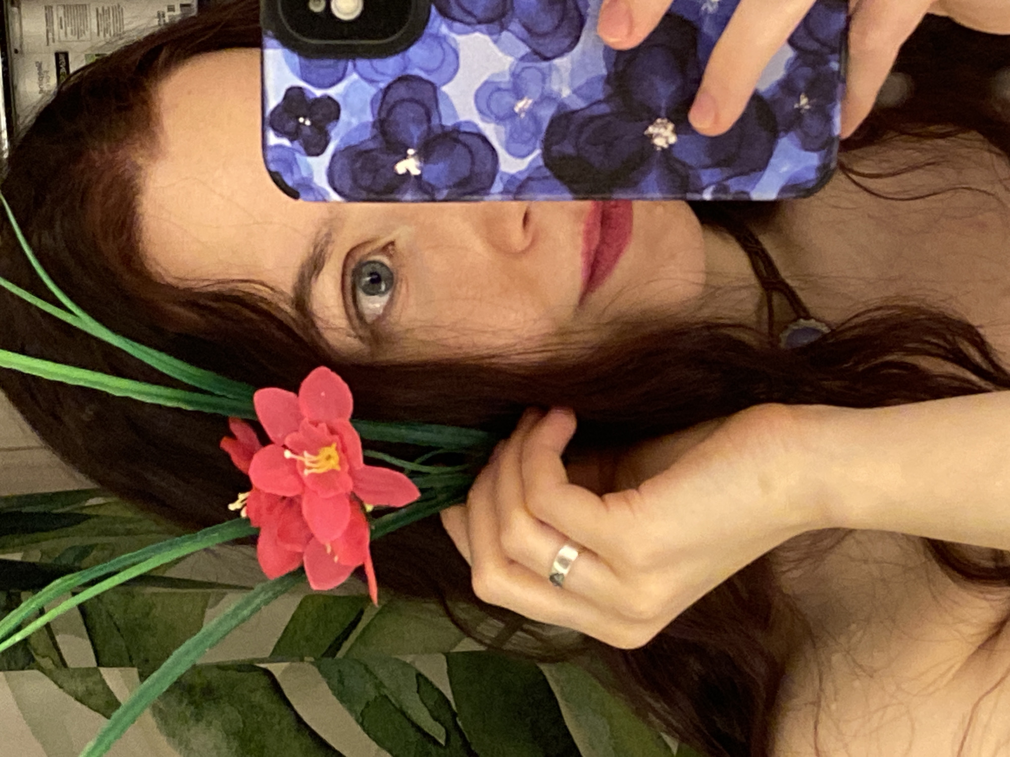 A woman is holding a cell phone in front of her face, obscuring part of it. The phone case has a blue and purple floral pattern. The woman has brown hair, blue eyes, and is wearing lipstick. She is holding a red flower with green stems and leaves. She is also wearing a ring on her finger. The background appears to be a bathroom with a green and white patterned wall. Without additional information it is not possible to verify if the location is in the city of Chaikovsky, Russia.