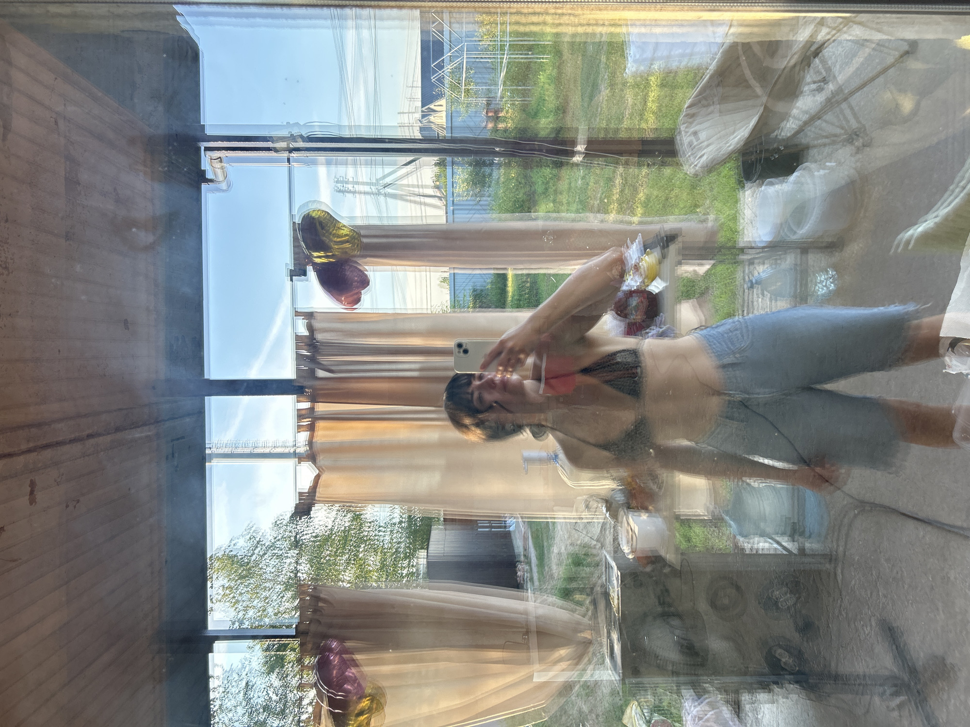 The image is a reflection in glass, appearing to be taken from inside a structure with a wooden ceiling and large glass panels, looking outwards. The reflection captures a young woman holding a smartphone, taking a selfie. She is wearing a dark patterned bikini top and light blue denim shorts. The background visible through the glass and in the reflection shows a suburban or rural environment, with green grass, trees, fences, and houses. There are also balloons, curtains, various objects on tables and shelves, and what appears to be a large water cooler bottle. The scene suggests a casual, outdoor setting possibly in a backyard or porch area, in the city of Pribrezhnyi, Russia, given the context provided. The lighting indicates it is daytime, possibly late afternoon with the sun casting shadows.