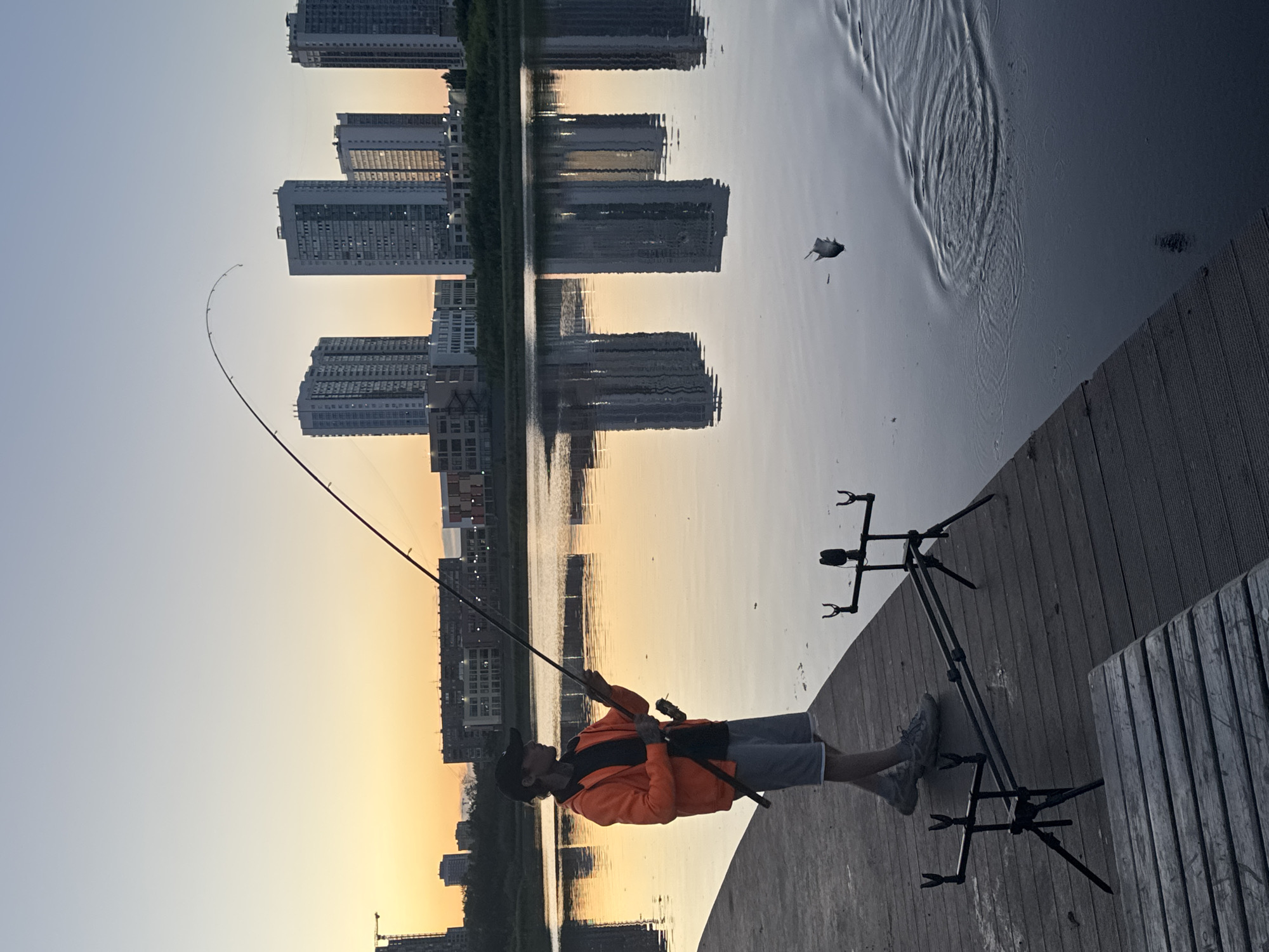 In Yekaterinburg, Russia, at sunset, a young man stands on a wooden pier, fishing. He is wearing an orange jacket, a dark t-shirt, grey shorts, a black baseball cap, and grey sneakers. He holds a fishing rod, with the line taut and curving towards the water, suggesting a catch. Beside him on the pier are two fishing rod holders. The water reflects the pale sky and the silhouettes of tall apartment buildings lining the opposite bank. In the water, a fish is airborne, mid-jump, with ripples spreading around it, indicating recent activity. The overall scene captures a moment of angling activity by the water in an urban setting during twilight.