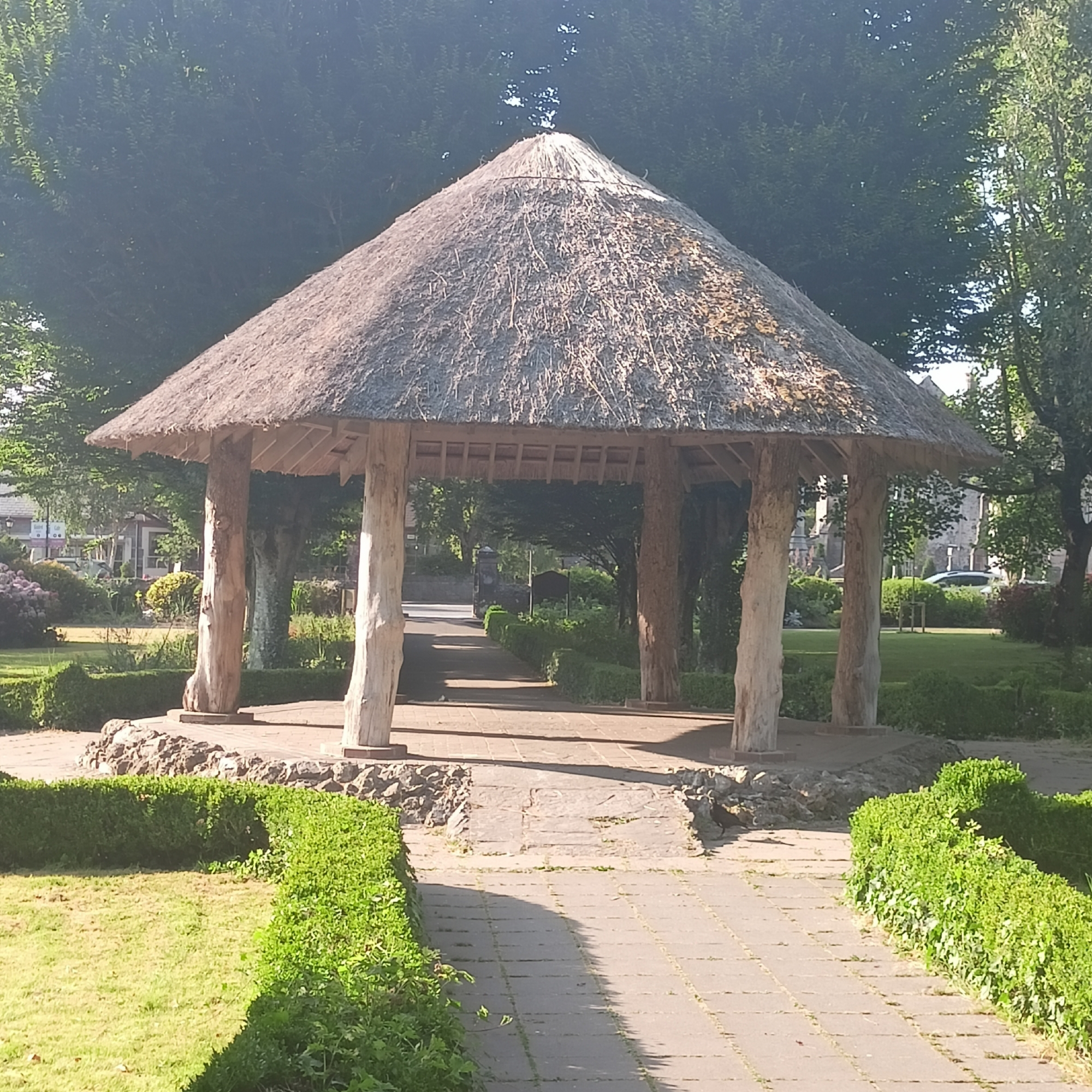The image depicts a thatched-roof gazebo with four large, rough-hewn wooden pillars, located in a park-like setting. The gazebo's roof is made of dry straw or reeds, supported by visible wooden beams. A paved pathway leads to and through the gazebo, bordered by meticulously trimmed green hedges on either side. Beyond the gazebo, the pathway continues, flanked by more hedges and green grass. In the background, mature trees with lush foliage create a dense canopy, with glimpses of sunlight filtering through. Further in the distance, some buildings and a car are visible, suggesting the park is near an urban or semi-urban area. The overall impression is a peaceful, well-maintained outdoor space. The specific location is identified as The Municipal District of Adare — Rathkeale, Ireland.