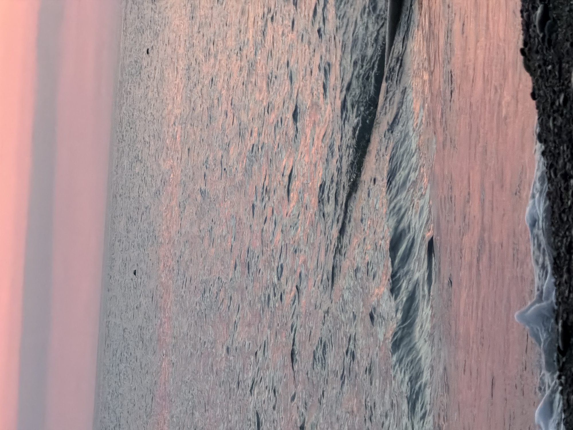 A tranquil seascape at dusk. The sky is a soft gradient of pink and lavender, reflecting gently on the calm ocean surface. Small, dark shapes, possibly birds or seals, dot the water in the distance. Gentle waves lap at a pebble-strewn shore in the foreground, where a small patch of white foam has gathered. The overall impression is one of peaceful serenity.