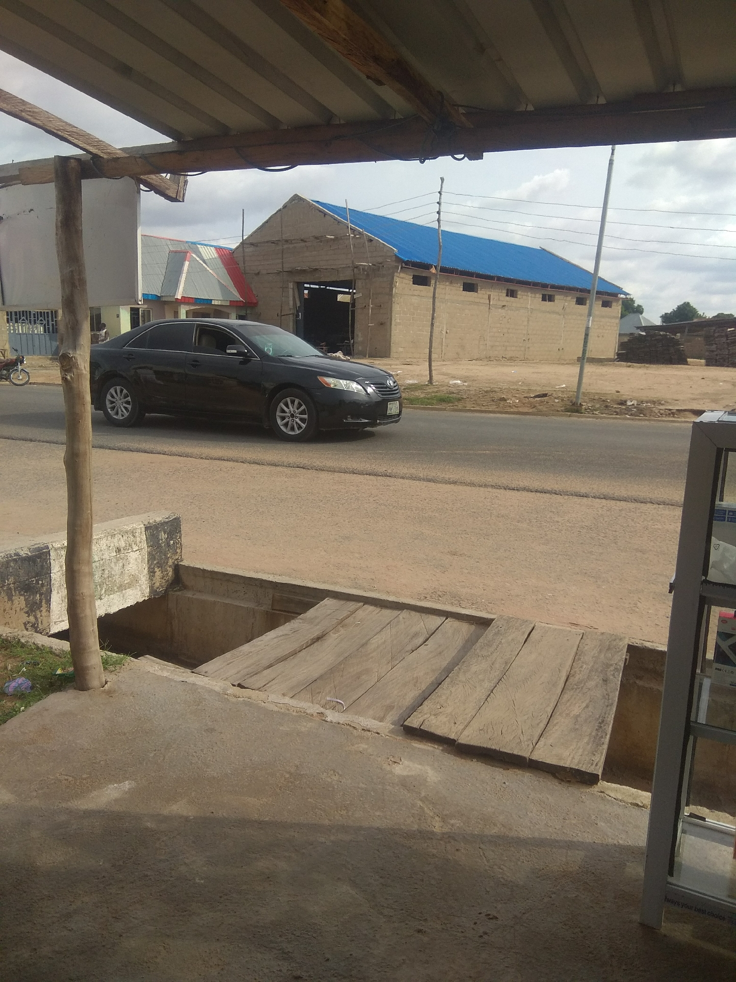 A black Toyota Camry is driving on a paved road in Jalingo, Nigeria. The car is in motion and is angled towards the right of the frame. In the background, there is a large building with a blue metal roof, and some smaller structures. There is also a wooden utility pole with wires extending from it. In the foreground, there is a concrete structure with wooden planks laid across it, possibly a makeshift bridge or cover over a drain. The ground is covered in dirt and gravel. The sky is overcast. There is no other visible activity or people in the frame.