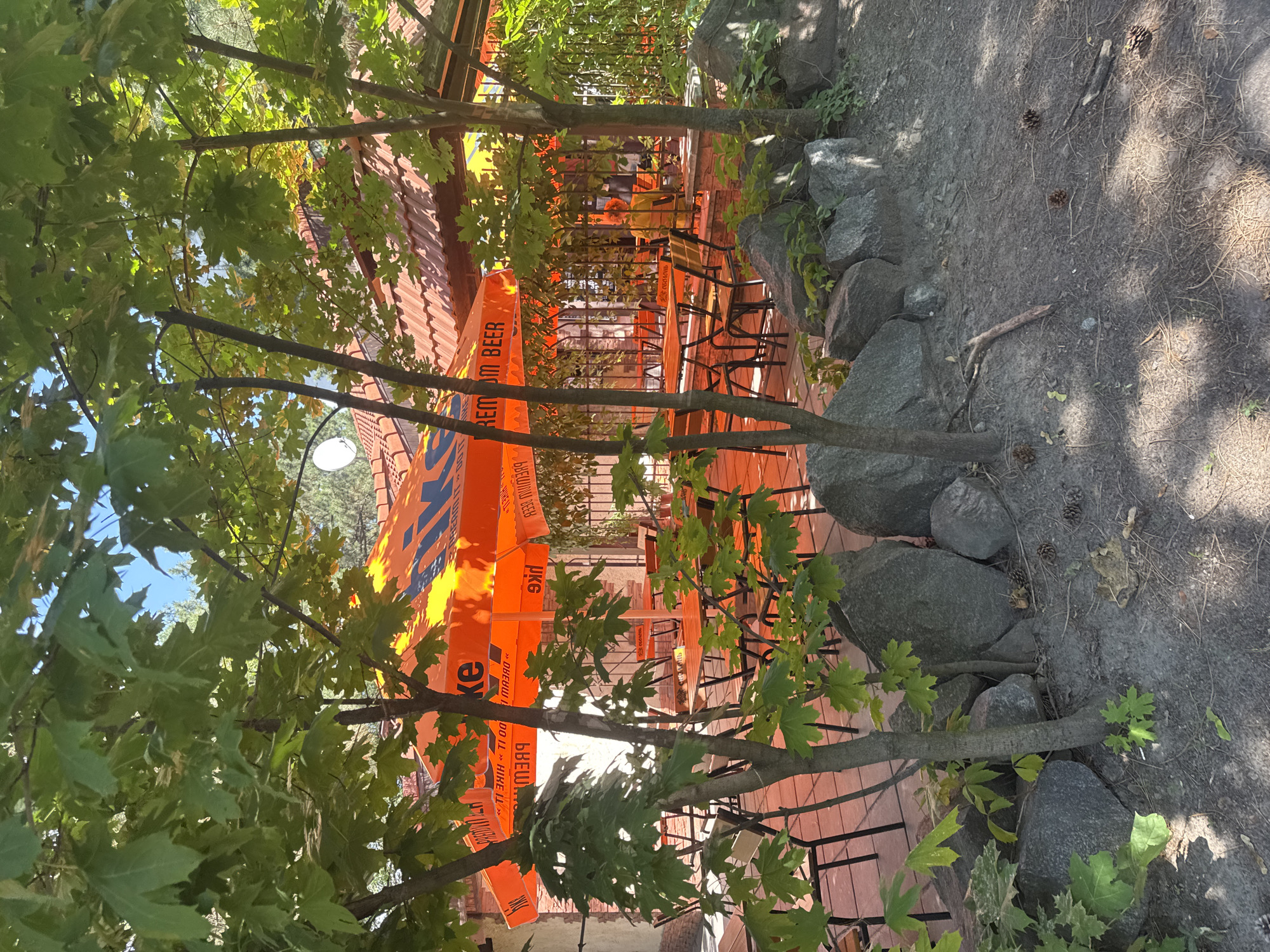 The image shows an outdoor seating area of a cafe or restaurant. Large orange umbrellas with "PREMIUM BEER" printed on them provide shade over several tables and chairs arranged on a tiled patio. The area is surrounded by trees with green leaves and a rock wall. In the background, a building with a red-tiled roof is visible. One person, wearing a yellow outfit, is seated at a table in the background. The scene is located in the city of Horishni Plavni, Ukraine.