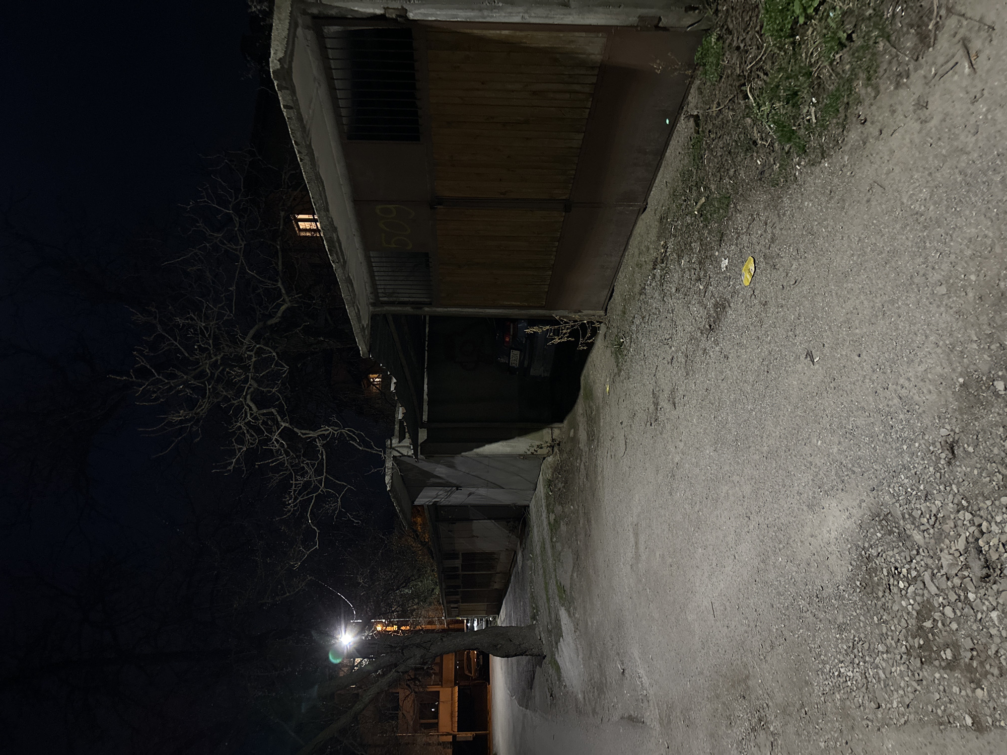 A nighttime scene depicts an unpaved dirt and gravel alleyway or access road in Sofia, Bulgaria, flanked by a row of garage structures. The ground in the foreground is uneven, comprised of light-colored dirt and various sized stones, with sparse green vegetation along the right edge. A single crumpled yellow item, possibly a wrapper, rests on the ground near the first garage structure.
On the right side of the image, a series of connected, single-story garage units are visible. The closest complete garage features a brown wooden gate for its lower section and dark metal bars for its upper section, surmounted by a concrete overhang. The number "509" is painted in yellow on the concrete beam above this gate. To the immediate left of this garage, another garage opening is partially visible, revealing the rear of a dark-colored passenger vehicle. The vehicle's license plate, "C 3536 KK", indicates its registration in Sofia, Bulgaria.
On the left side of the alley, a tall, bare-branched tree stands next to a street light pole. The street light emits a bright, white-green illumination, casting light and shadows across the scene. Further in the background on the left, additional residential or commercial structures are partially visible, one with illuminated windows. The sky is dark and clear, showing no celestial bodies. A distant building beyond the garages on the right also displays a single illuminated window. No individuals are present in the scene.