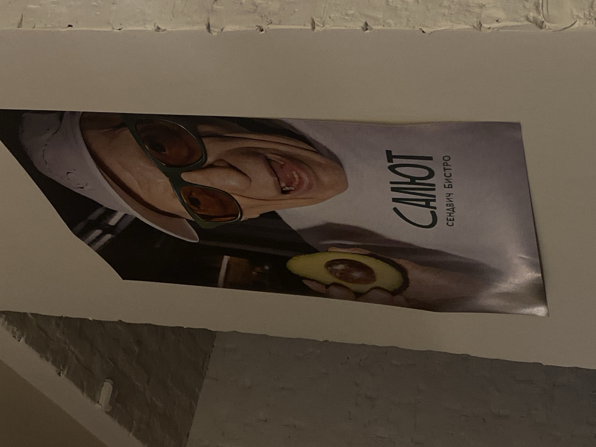 The image depicts a poster advertising a restaurant in Krasnoyarsk, Russia. The poster features a person wearing sunglasses and a chef's hat, holding half an avocado. The text on the poster reads "Салют Сендвіч Бістро," indicating the name of the establishment is "Salyut Sandwich Bistro." The poster is affixed to a white wall.