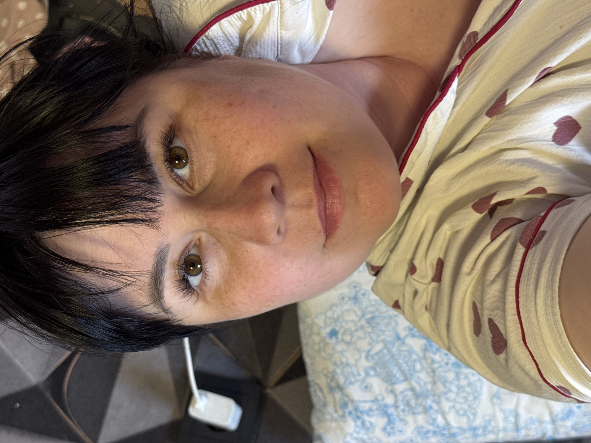 A selfie-style image depicts a woman with dark hair and bangs, fair skin with freckles, and hazel eyes. She is wearing a white top with red trim and maroon heart shapes. She is lying on a surface with patterned bedding and a tiled floor. A white object with a cord is visible near her hair. Zav'yalovo, Russia, is not identifiable from the visual information provided.