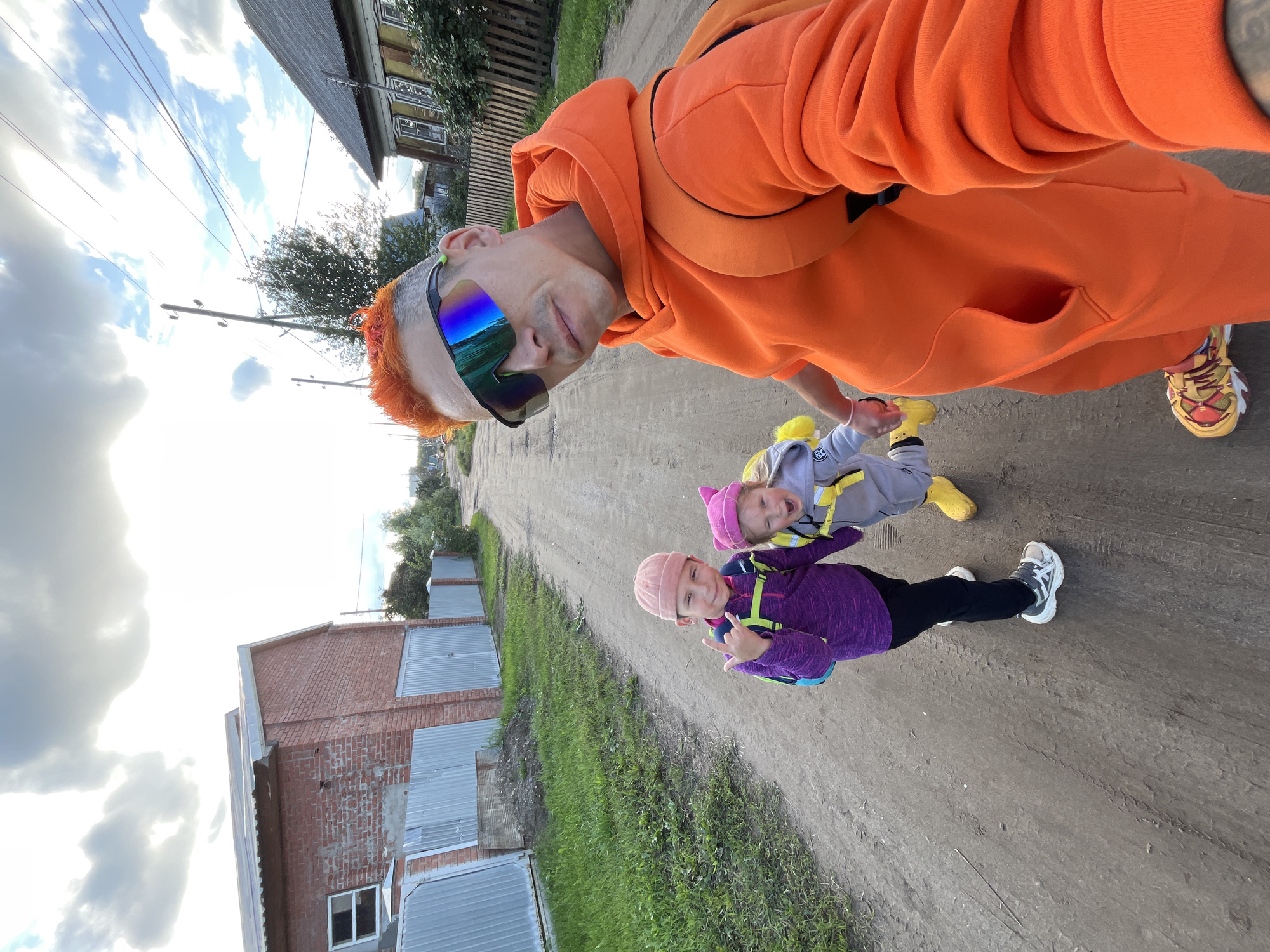 In a selfie taken outdoors, a man with brightly dyed orange hair and reflective sunglasses, wearing an orange hooded sweatshirt and sweatpants, stands with two children on a dirt road. The older child, on the left, wears a purple jacket, black pants, and a pink hat, and makes a rock-on hand gesture. The younger child, in the center, wears a gray outfit, a pink hat, and yellow boots, and appears to be crying or yelling. The man is holding the younger child's hand. In the background, there are houses with wooden fences, a brick building with a metal fence, and utility poles with wires, suggesting a rural or suburban setting. The sky is cloudy, and the overall environment appears to be a street in Chaikovsky, Russia, given the architectural style of the houses and the presence of a dirt road.