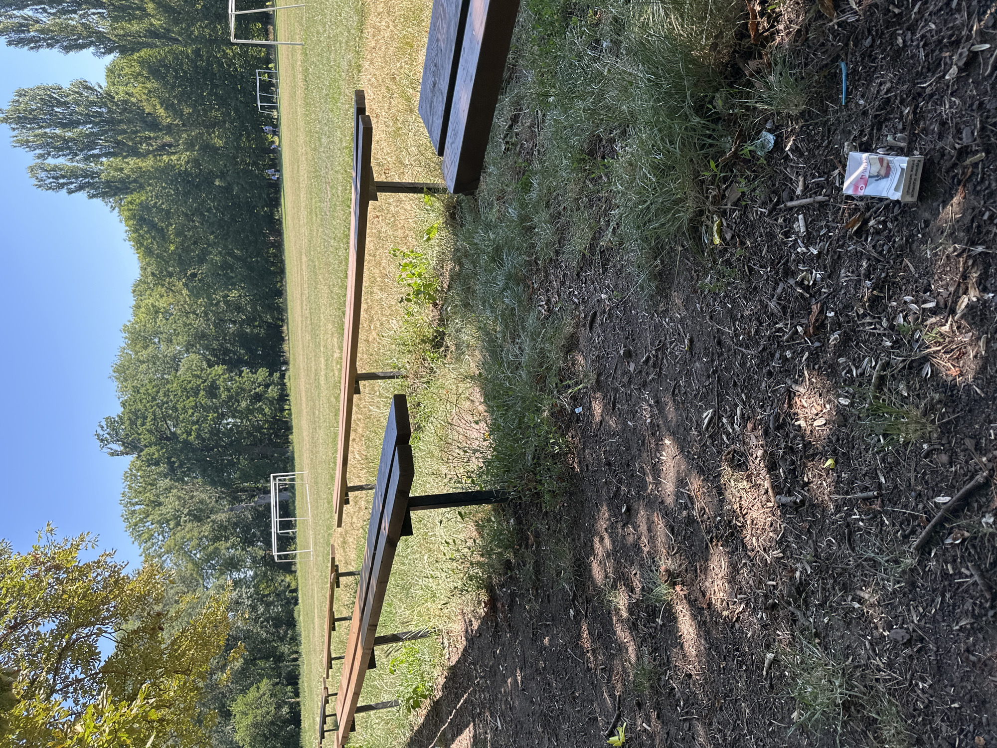 In Makiv, Ukraine, a daytime, eye-level shot depicts a scene on what appears to be a sports field. Wooden bleachers, weathered and showing signs of use, are positioned in the foreground. The ground is a mix of dark soil and dry grass, with scattered debris including a crumpled cigarette pack. In the background, a soccer goal is visible on a grassy field, bordered by a dense line of trees under a clear blue sky. Several indistinct figures can be seen in the middle ground, suggesting light activity on the field. The overall impression is one of a public outdoor space on a bright, warm day.