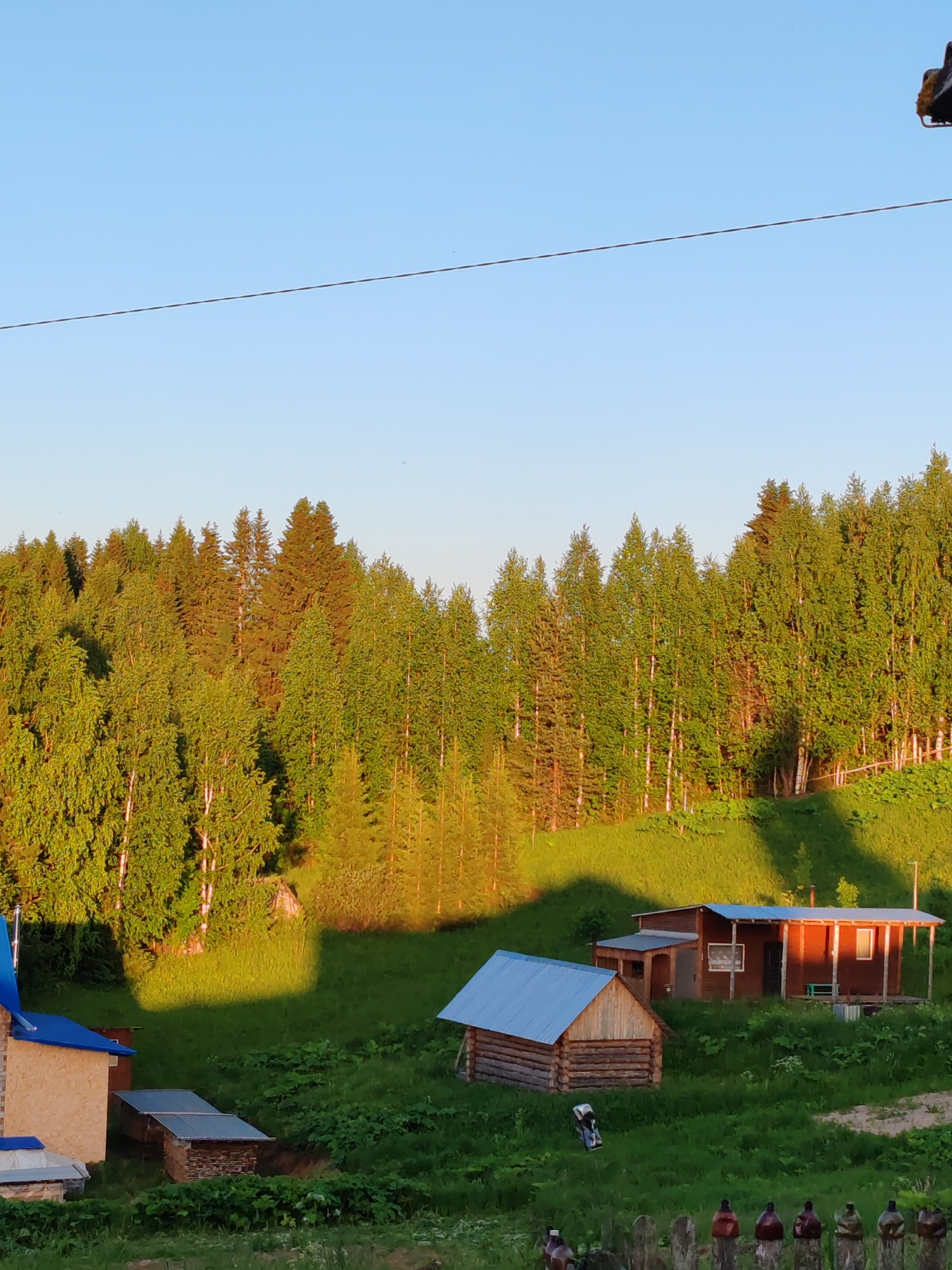 This detailed image captures a rural landscape in Чухлэм, Russia, under clear, bright conditions, likely during sunrise or sunset, indicated by the golden light and long shadows.
The upper third of the frame consists of a cloudless, light blue sky. A thin, dark, horizontal wire or power line spans across the sky, slightly above the midpoint of the sky segment.
The middle ground is dominated by a dense forest covering a gently sloping hillside. The forest comprises a mix of deciduous trees, primarily birches with their distinctive white trunks and bright green foliage, and coniferous trees, which appear darker green and more densely packed. The sun illuminates the tops and front-facing sides of the trees with a warm, golden glow, creating a strong contrast with the shadowed areas within the forest canopy.
Below the forest line, the hillside is covered in lush green grass and scattered low-lying vegetation. Large, dark shadows are cast across this grassy slope from unseen objects or the forest itself, creating distinct patches of light and shade.
Several structures are present in the lower half of the image. On the far left, a light-colored building with a bright blue pitched roof is partially visible. Adjacent to it, a smaller structure with a corrugated metal roof is partially embedded in the ground. In the center-right foreground, a small, rustic log cabin with a light blue corrugated metal roof stands on the grassy slope. To its right and slightly behind it, a larger, dark reddish-brown wooden house with a light blue corrugated metal roof features a covered porch extending along its front facade, with visible windows and a door. A white, irregularly shaped object lies on the grass in front of the log cabin.
In the bottom right corner, a wooden fence is partially visible, with several plastic bottles, varying in color from dark brown to camouflage green, placed upright on the fence posts. No human figures or animals are present in the scene.