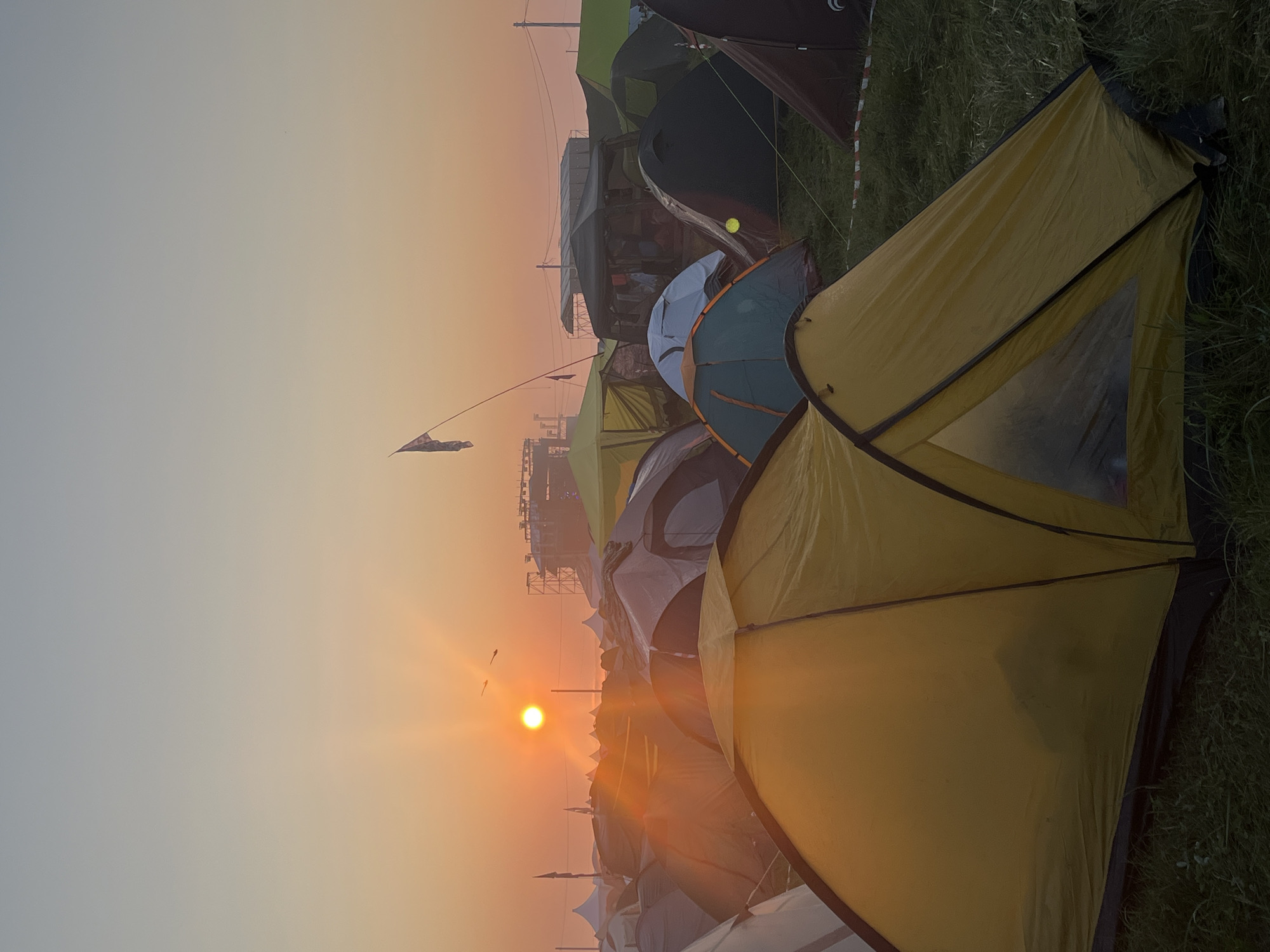 The image depicts a sunrise over a camping area, likely a festival or event, filled with numerous tents. The sun, a bright orange orb, is low on the horizon, casting a warm glow across the scene and creating lens flares. The sky transitions from a soft orange near the sun to a pale blue at the top. A stage structure or set of lights is visible in the distance, suggesting the presence of a large event. A flag on a pole flutters in the breeze. The foreground is dominated by a yellow tent, with other tents of various colors and sizes scattered throughout the middle ground and background. The ground is covered in grass. There are no people visible in the image. The location is identified as Surakovo, Russia.