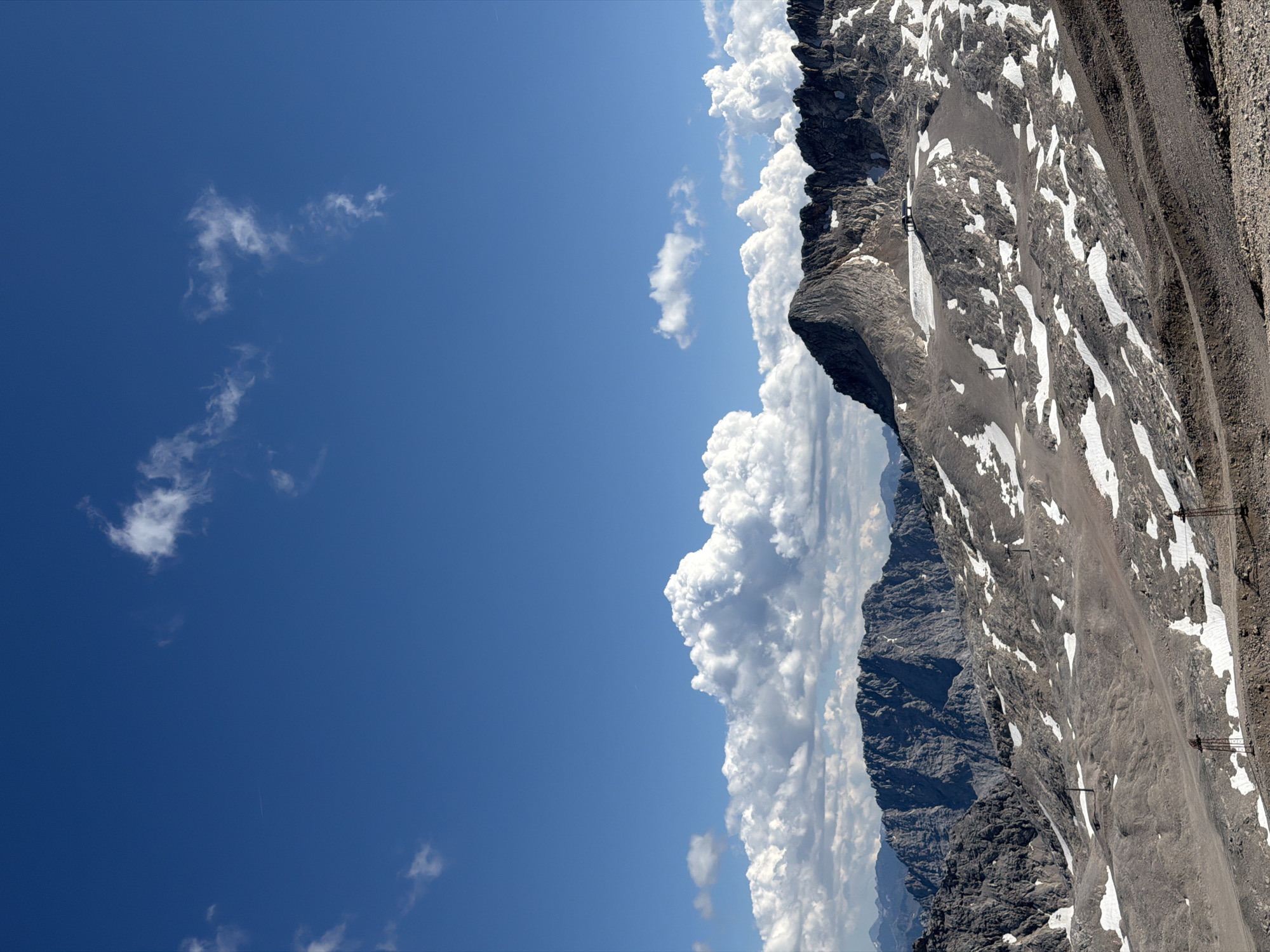 The image depicts a mountainous landscape under a bright blue sky dotted with scattered white clouds. The foreground features a rocky, barren terrain with patches of snow. In the mid-ground, a range of jagged, dark gray mountains rises, some peaks still covered with snow. Several poles, likely associated with ski lifts or infrastructure, are visible on the slopes. The background shows a further chain of mountains fading into the distance. The scene is devoid of any people. The location is the Zugspitze massif, the highest point in Germany, situated on the border between Germany and Austria, near the Bavarian town of Garmisch-Partenkirchen.
