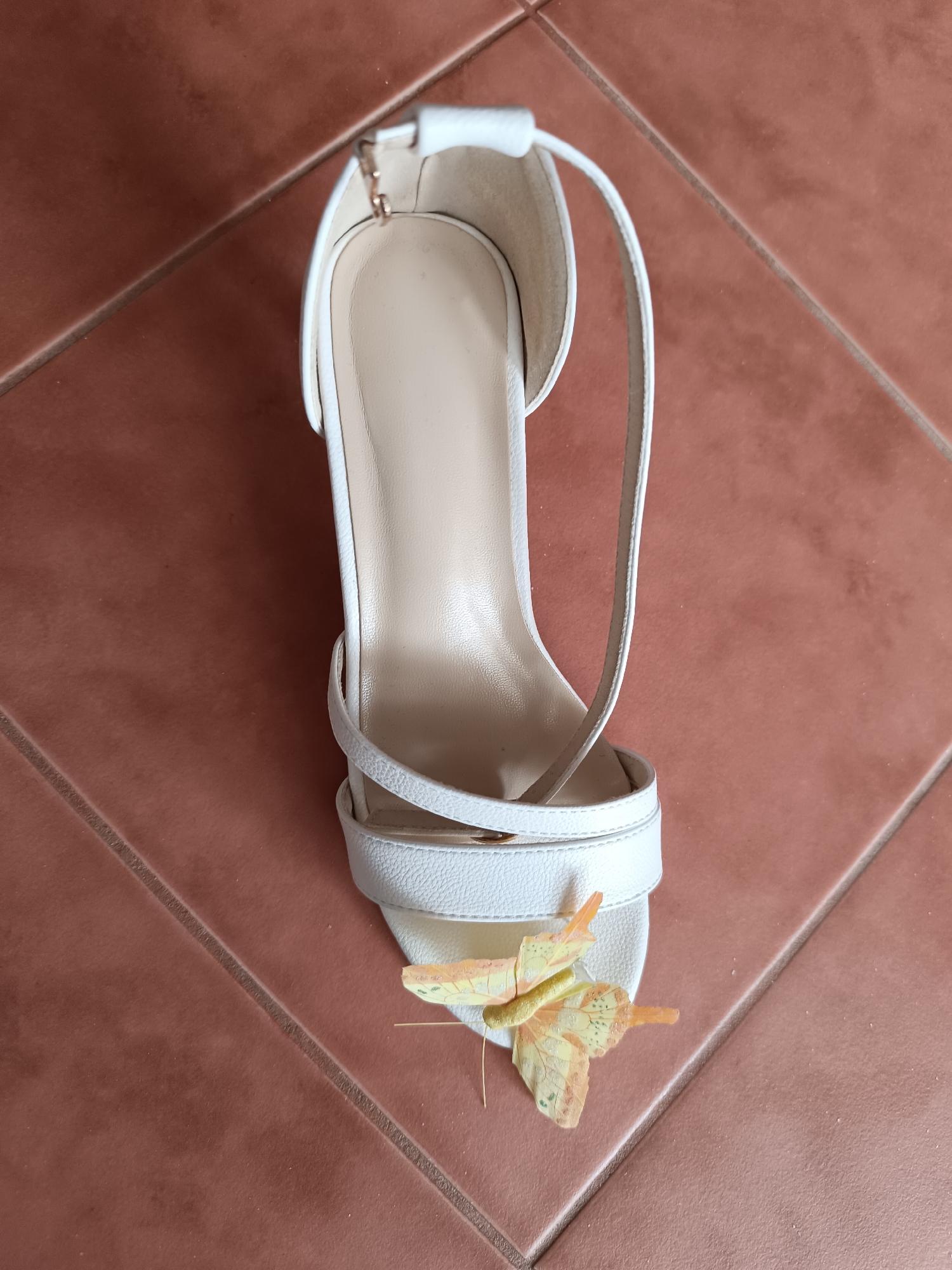 A white high-heeled sandal with a delicate butterfly decoration is placed on a floor of reddish-brown tiles. The sandal has a strap around the ankle and straps across the toes. The butterfly is light yellow with orange accents. The floor tiles are arranged in a diagonal pattern. The image appears to have been taken indoors, possibly in Ternopil, Ukraine, though this cannot be definitively confirmed from the visual evidence alone. There are no people or actions depicted in the image.