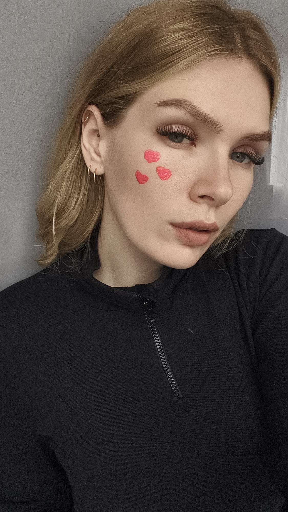 A woman is shown in a close-up studio shot against a gray wall. She has fair skin, blonde hair, and light-colored eyes with long eyelashes. There are three pink heart-shaped designs painted on her left cheek. She is wearing a black zip-up top. There is no information available to ascertain the location as Русский Вожой, Russia.