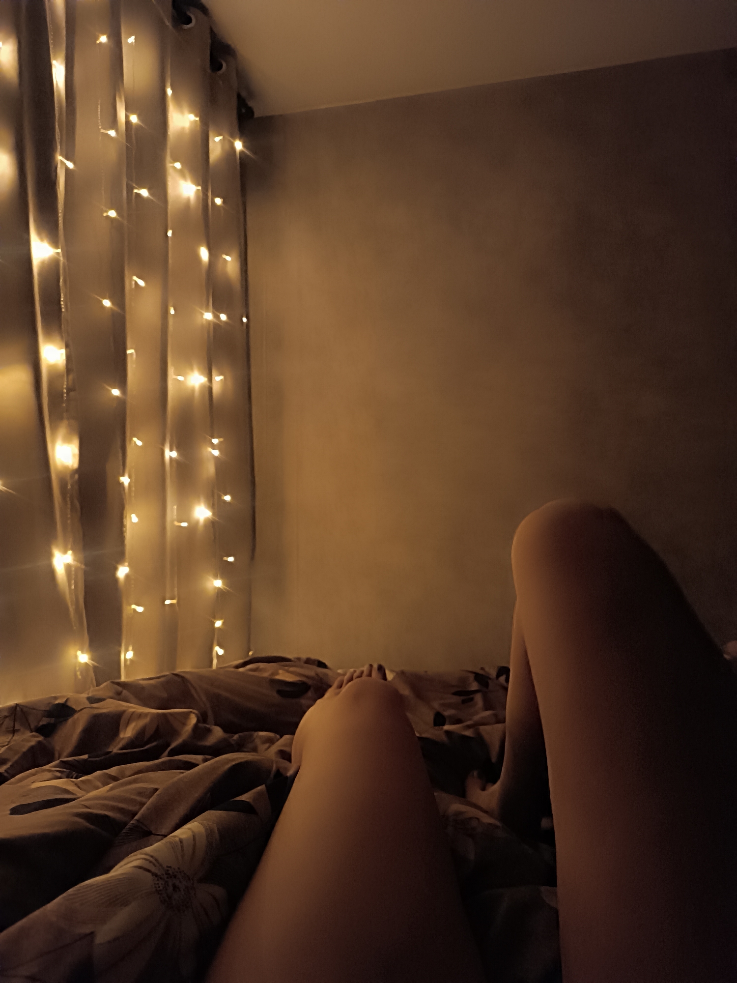 The image shows a view from someone lying in bed. The person's legs are visible, with one leg closer to the camera. The bed is covered with a patterned duvet. To the left of the bed are curtains adorned with string lights. The room has a dimly lit, warm tone. The location is unknown.