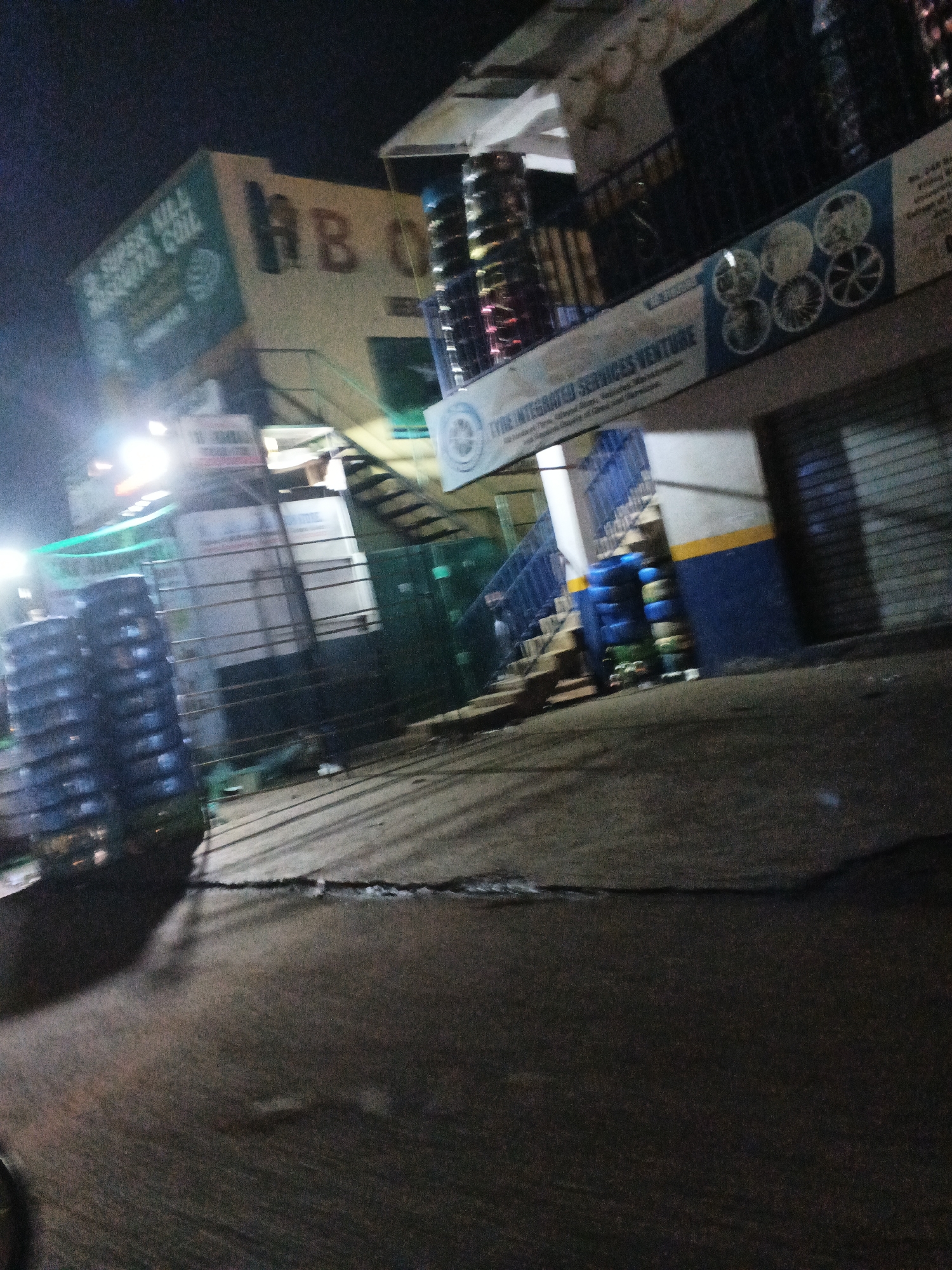 The image depicts a nighttime street scene in Jos, Nigeria, likely in front of commercial establishments. A prominent building, painted a light cream color, displays the signage "HB O" in large, dark letters. To the left of this, a green sign reads "SUPER HIL AUTO COIL". Above the "HB O" sign, a narrow staircase leads upwards. To the right of the main building, a banner advertises "TYRE INTEGRATED SERVICES VENTURE" and shows circular images of car wheels. Stacked tires, colored blue and green, are visible near the stairs. In the foreground, stacks of dark-colored, possibly plastic, crates or containers are arranged, and the ground appears to be paved with a rough asphalt surface, showing some cracking. Artificial lighting, including a bright light on the left, illuminates the scene. The overall impression is one of a busy commercial area at night, with goods and services being offered.