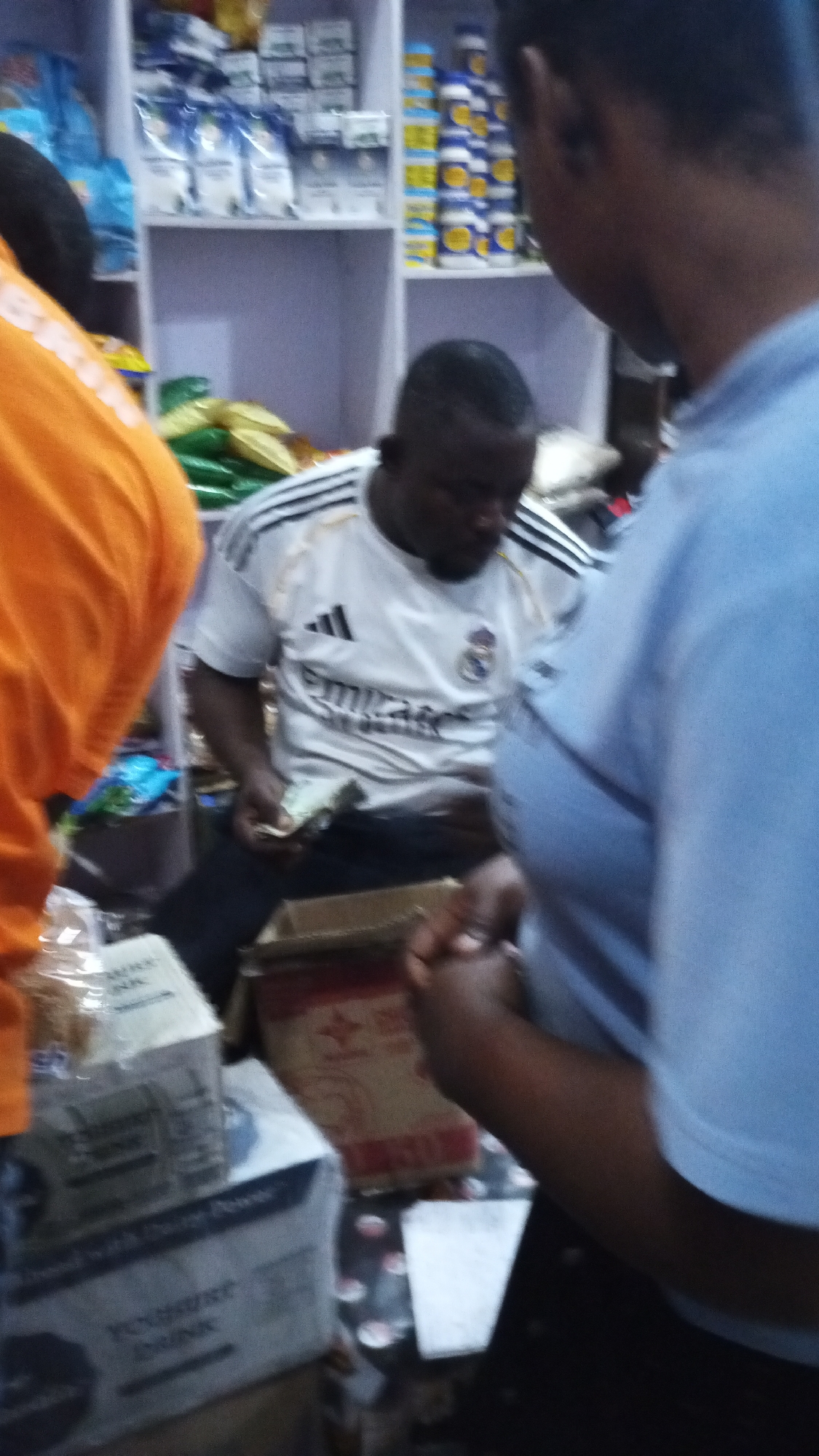 The image captures an indoor scene, likely within a small store or shop in Adankolo, Nigeria. Three individuals are visible. On the left, a person wearing an orange shirt is partially seen from the back, with some faint text, possibly "BRT," on their shoulder. In the center, a man wearing a white Adidas shirt with "Emirates" and the Real Madrid crest is seated, seemingly engrossed in handling a stack of banknotes. His head is slightly bowed, indicating focus. On the right, the back of a person's head and torso, clad in a light blue top, are visible, suggesting they are standing near the seated man.
The background features white shelving units stocked with various packaged goods. On the upper shelves, there are numerous white bags or boxes, and to their right, rows of yellow and blue containers. A lower shelf or surface displays items in green and gold packaging. In the foreground, several boxes are present. On the left, a box is partially visible with the text "Yogurt Drink" clearly legible. Another open brown cardboard box is positioned between the seated man and the person in blue. The activity centers around a potential transaction or money handling within what appears to be a retail or supply setting. The general lighting suggests it is daytime indoors, with no external weather cues.