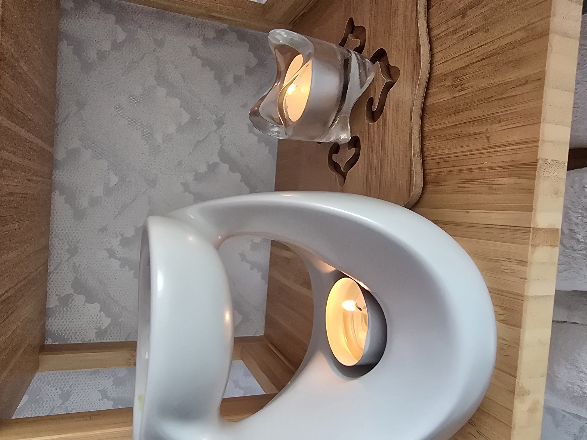 The image shows a close-up view of two lit tea light candles on a wooden shelf. One candle is inside a white ceramic oil diffuser, and the other is in a small clear glass votive holder. The background features a wall with a textured, light gray pattern and a wooden shelf above. The scene is indoors and appears to be in a residential setting, possibly a bathroom or spa area. There are no people present in the image. The location is not determinable from the image.