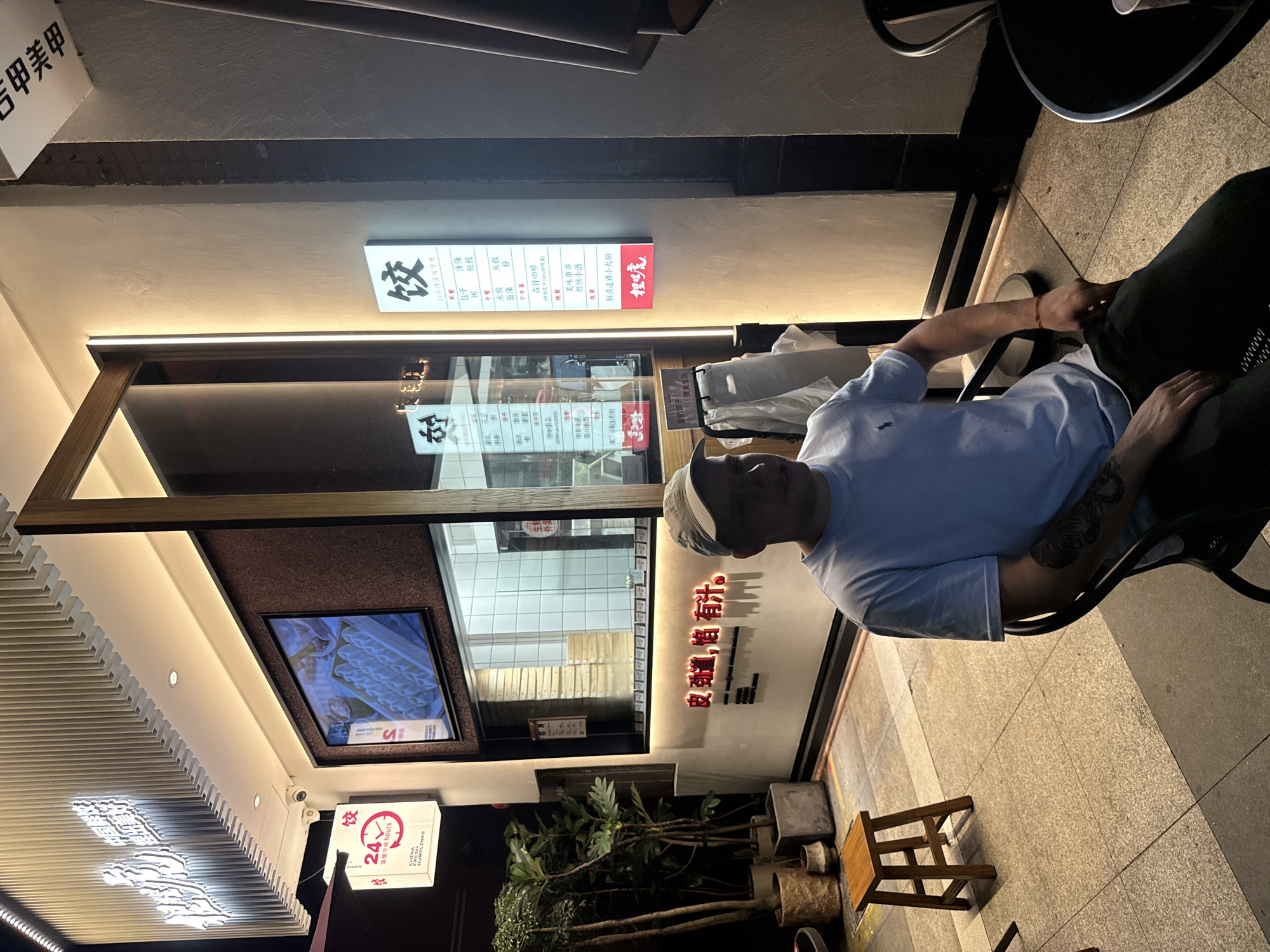 The image shows a man sitting at a table outside a restaurant. He is wearing a light blue t-shirt and a baseball cap. He has tattoos on his arm. The restaurant appears to be open, with a lit menu displayed on the wall and a TV screen showing food. The signage includes Chinese characters and indicates that the restaurant is open 24 hours and serves dumplings. There are also potted plants and a small wooden stool visible in the foreground. The scene is lit by artificial light, suggesting it is nighttime. The location is identified as Haizhu District, China. The overall mood is casual and relaxed.